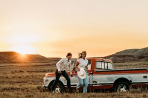 A couple shares a sunset moment on their vintage pickup truck in a golden prairie landscape.