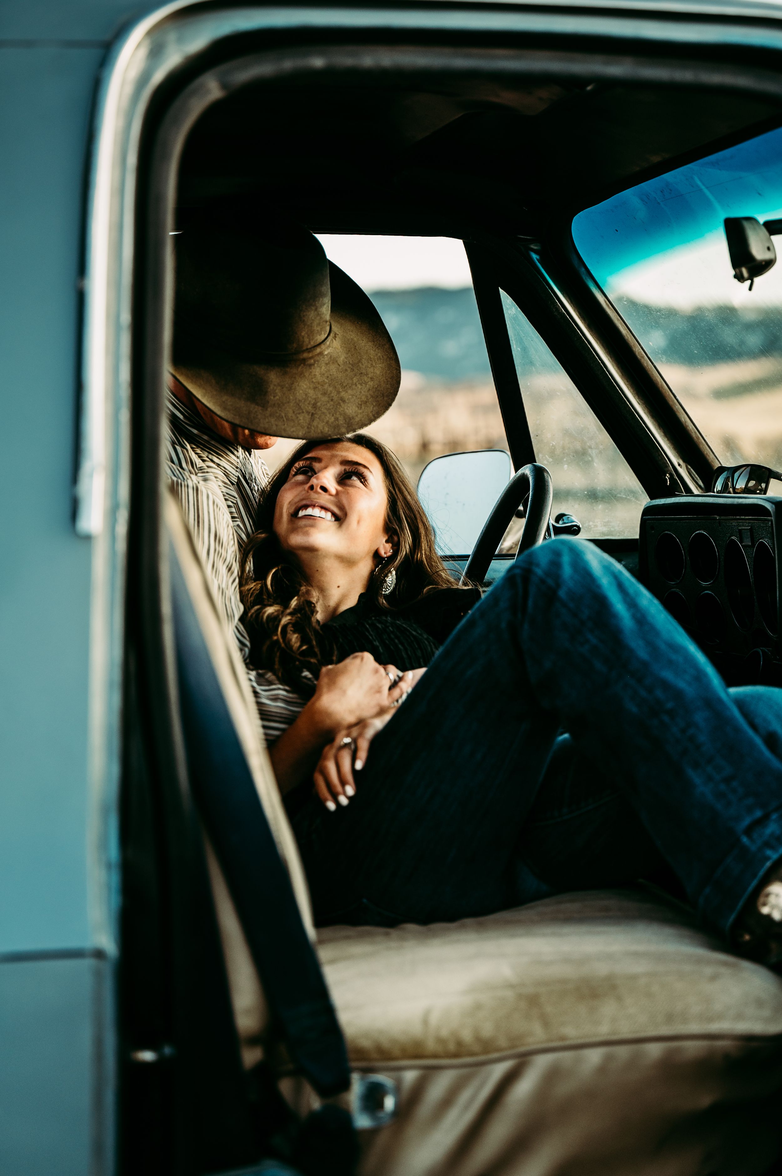 A laughing woman in jeans sits in a vintage truck cab while a person in a wide-brim hat leans in close.