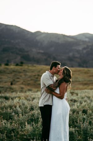 Couple sharing a romantic kiss in a scenic mountain meadow, woman in white dress, golden hour light.