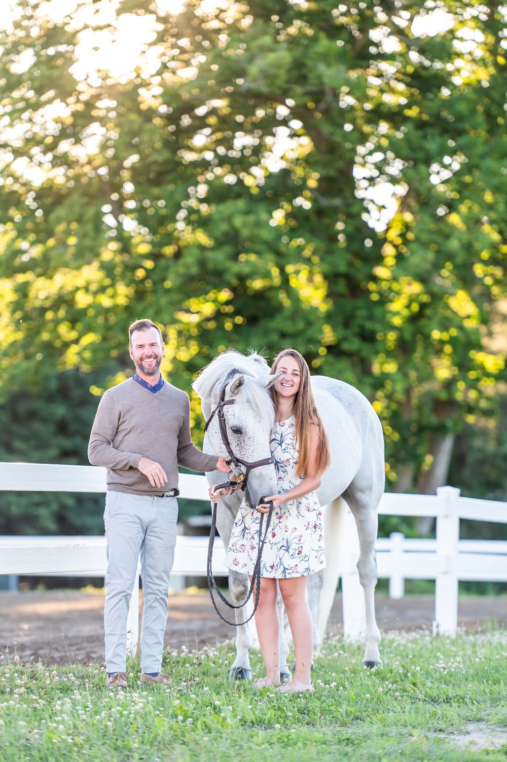 A couple stands with their white horse by a wooden fence in a sunlit pastoral setting.