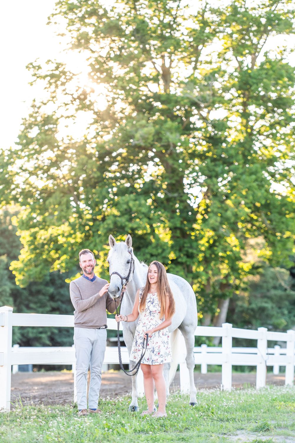 A couple poses with their white horse near a white fence at sunset in an outdoor equestrian setting.
