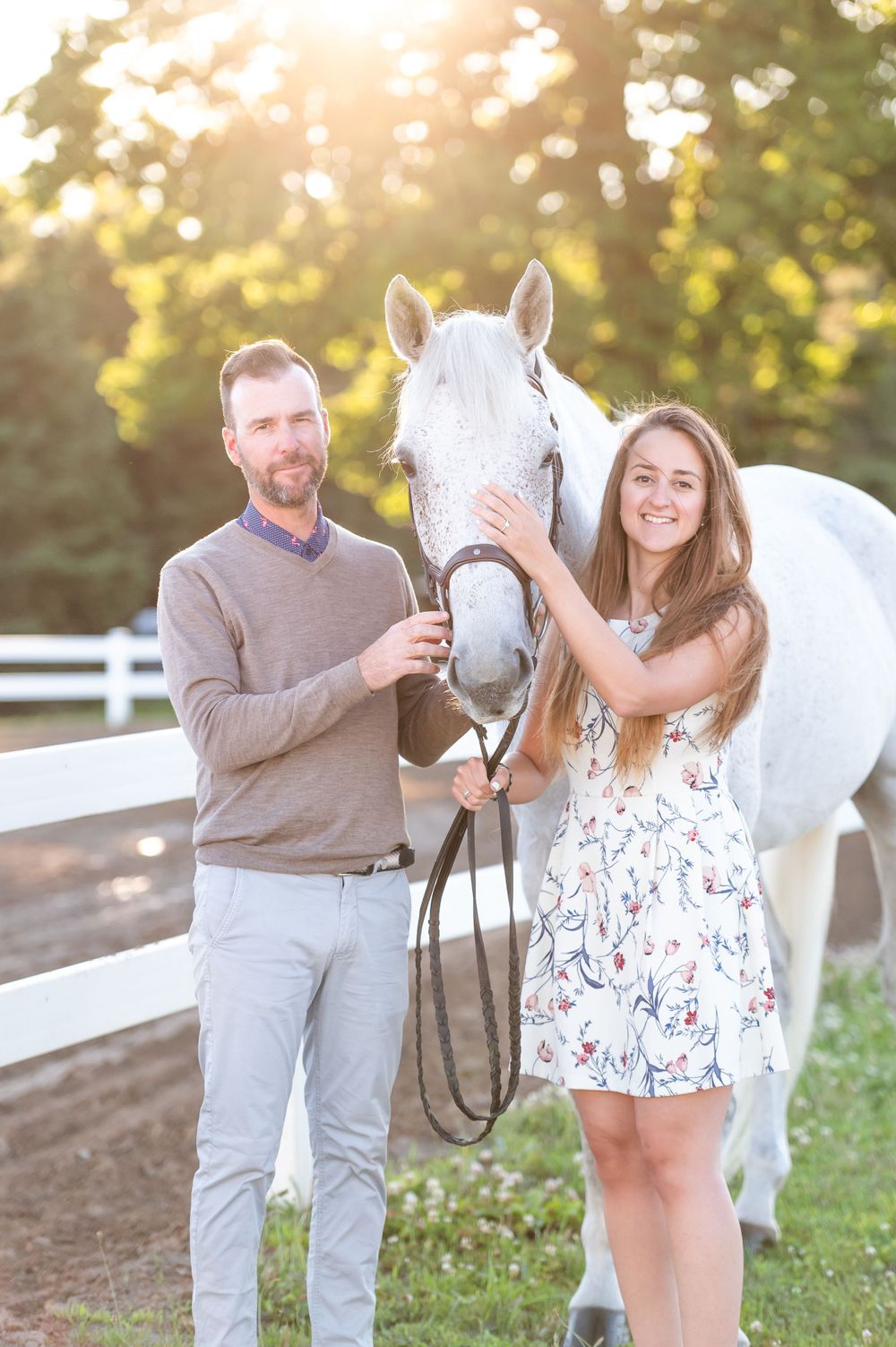 A couple poses with their beautiful white horse at sunset by a white fence on a farm.
