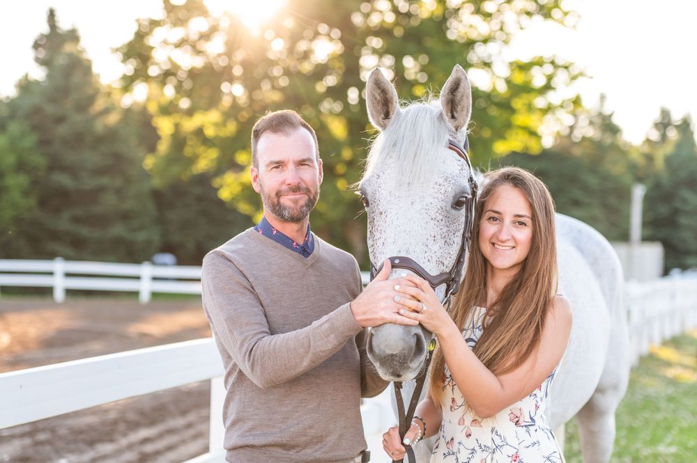 A couple poses outdoors with their white horse along a fence at sunset with warm golden lighting.