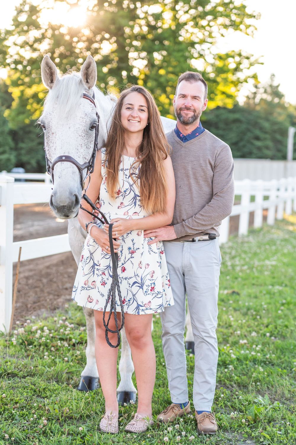 A couple stands with their beautiful white horse on a grassy field with white fencing in the background.