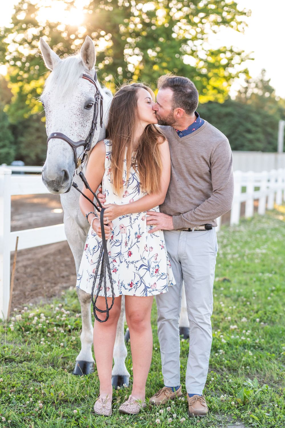 Romantic couple shares intimate moment with their white horse against backdrop of white fence during golden hour.