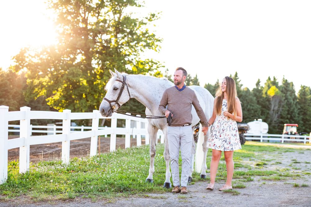 Couple walking with a white horse along white fence at sunset on farm property.