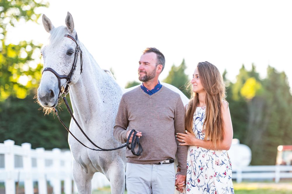 Couple standing with a beautiful white horse in front of white fence during golden sunset light.