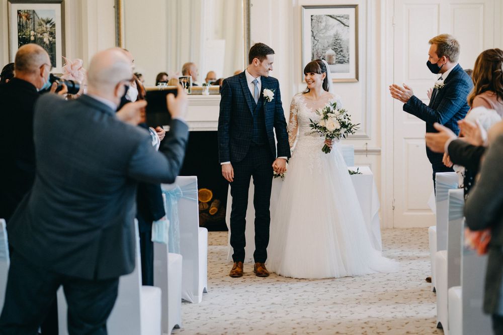 Wedding guests photograph a couple walking down the aisle at an elegant indoor ceremony venue.