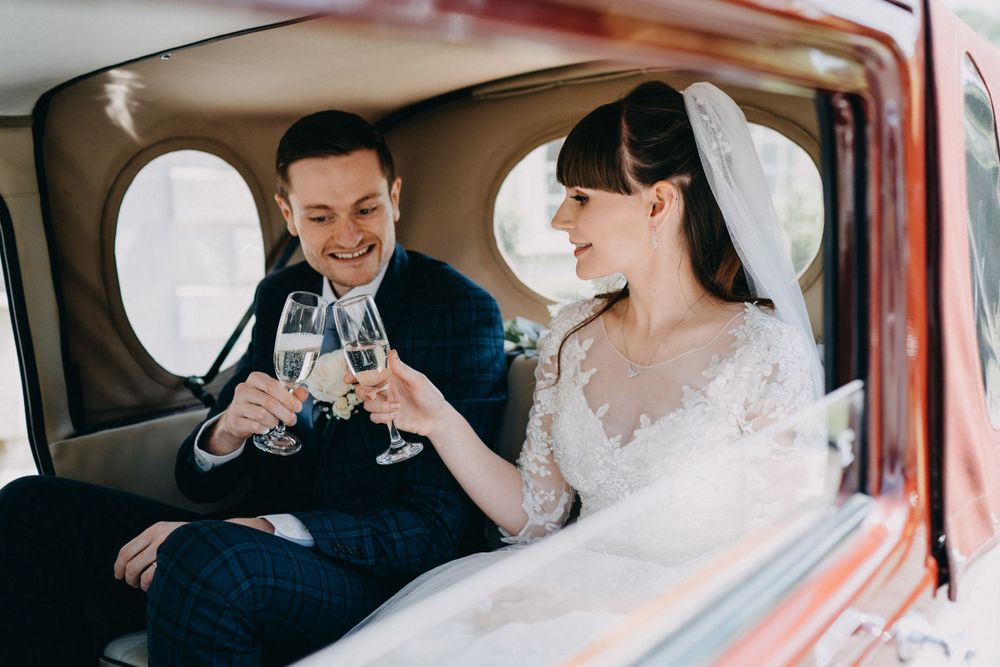 Couple toasting with champagne glasses inside vintage wedding car.