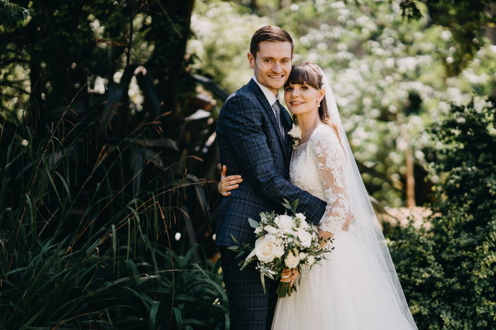 A romantic wedding portrait of a couple embracing outdoors with a white floral bouquet and lace dress details.