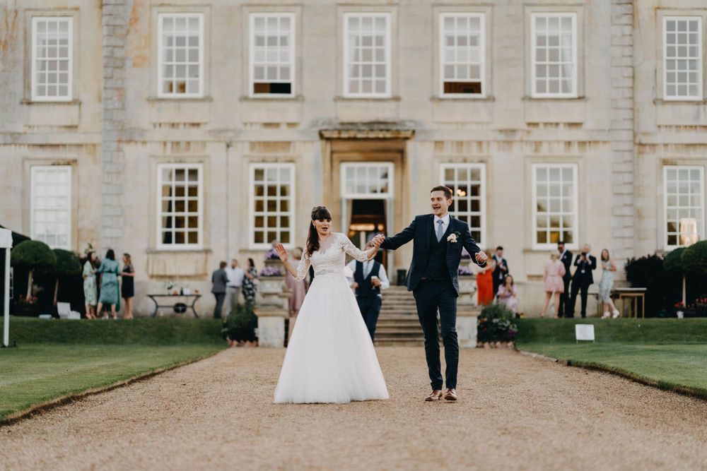 Elegant couple walking along gravel path towards historic mansion during wedding celebration at Harrowden Hall.