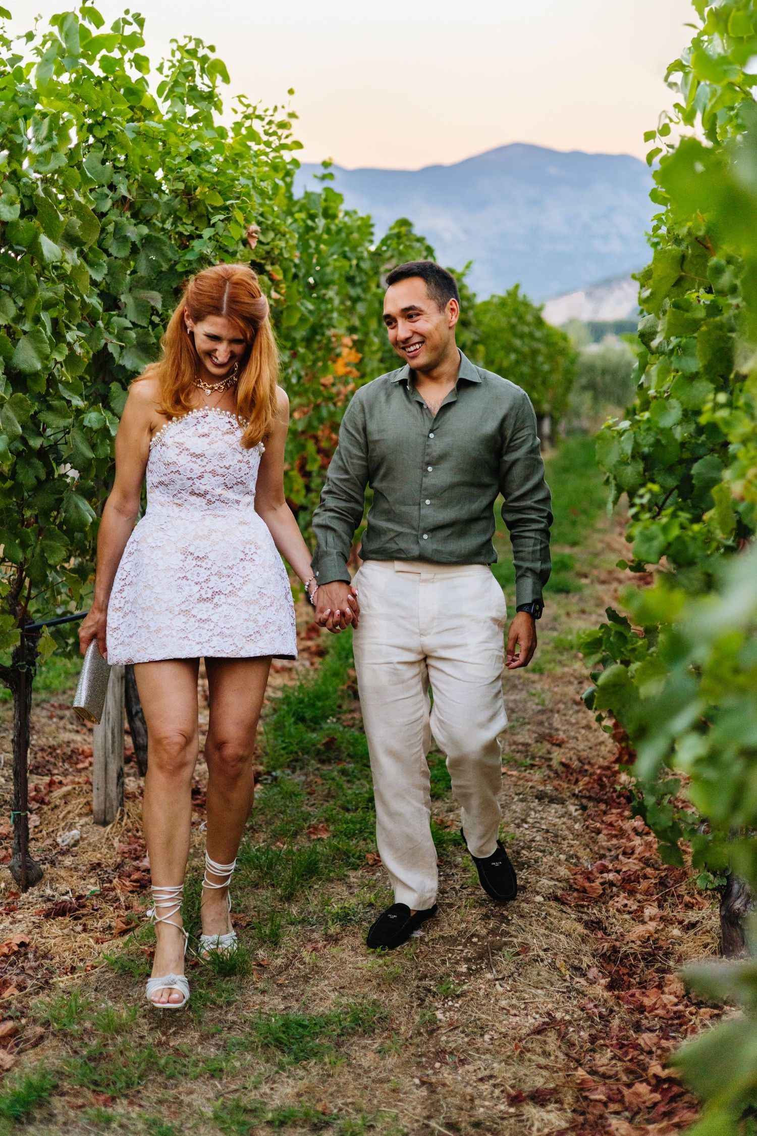 Happy couple holding hands while walking together through scenic vineyard rows at sunset with mountains in background.