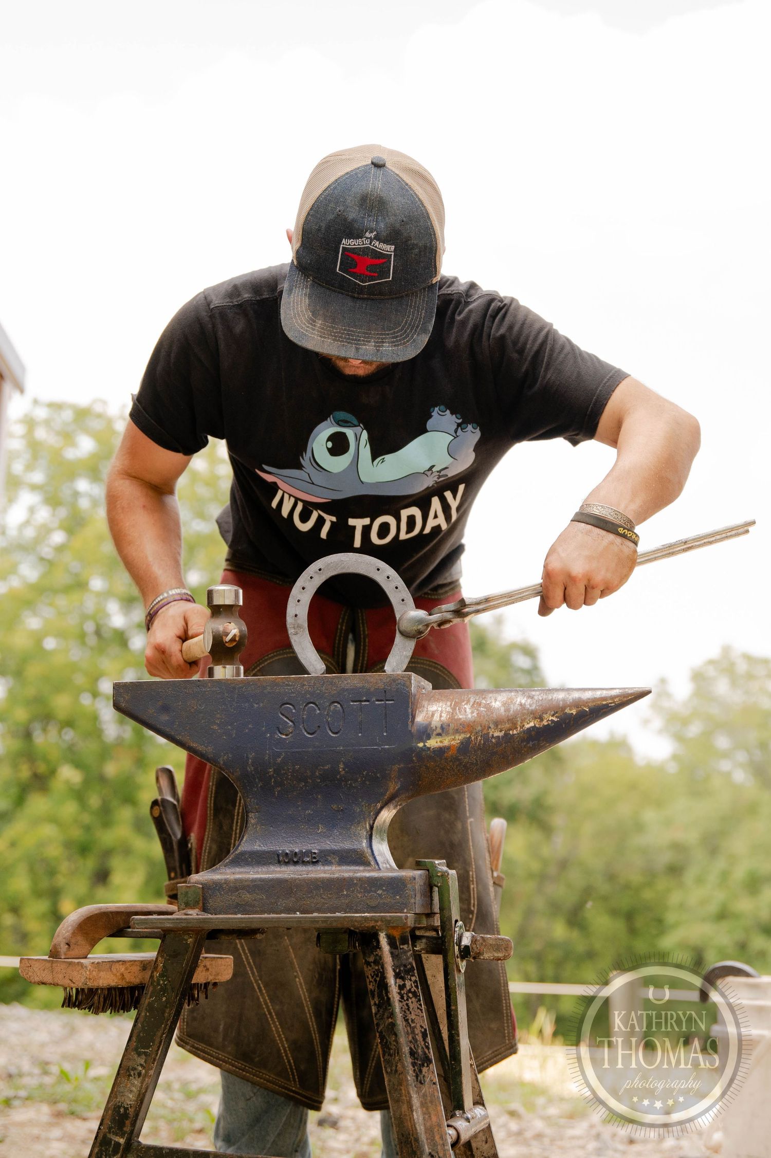 A farrier hammering hot metal on an anvil while wearing a black t-shirt and cap.