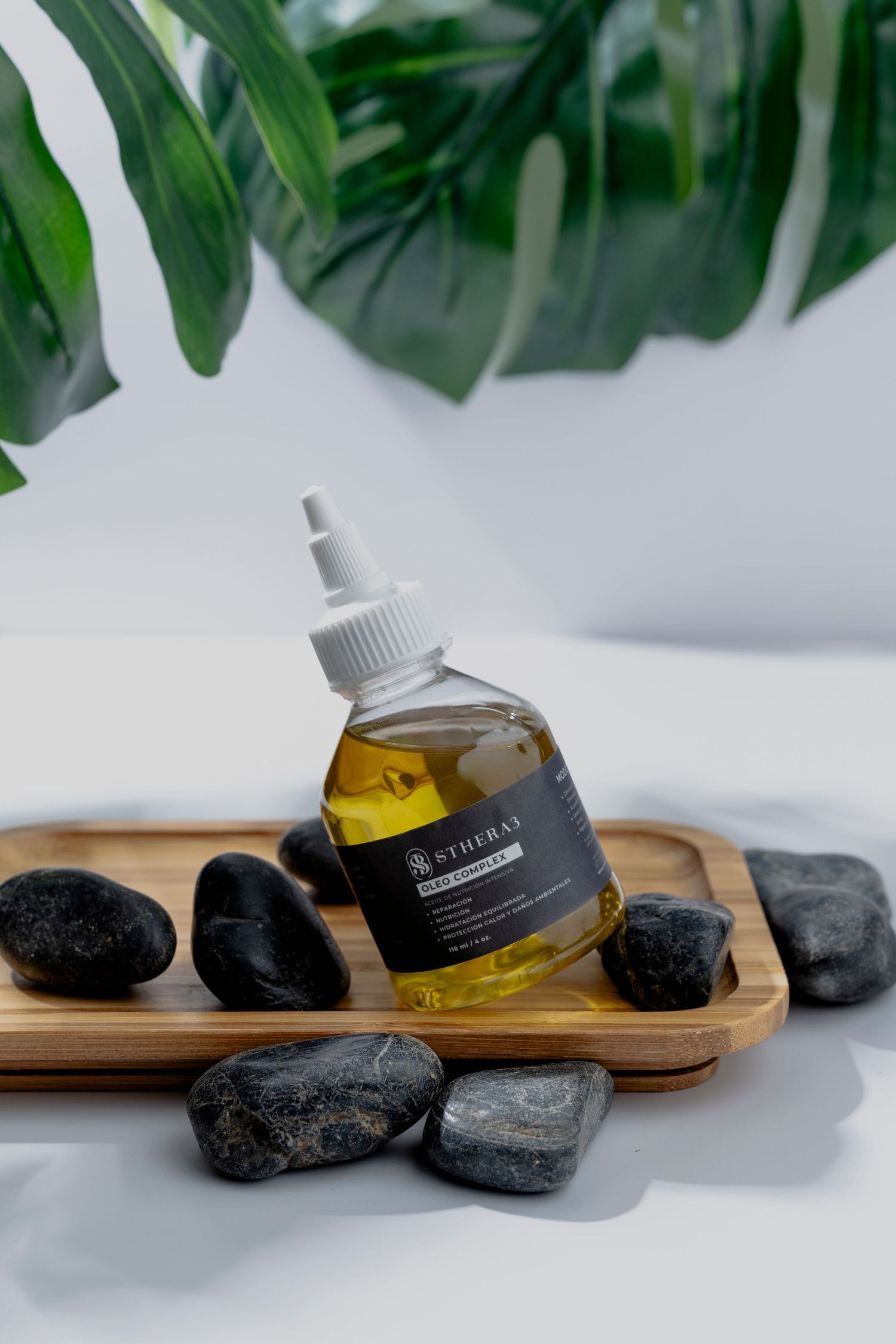 Aromatherapy oil bottle arranged with black stones and tropical leaves on wooden tray.