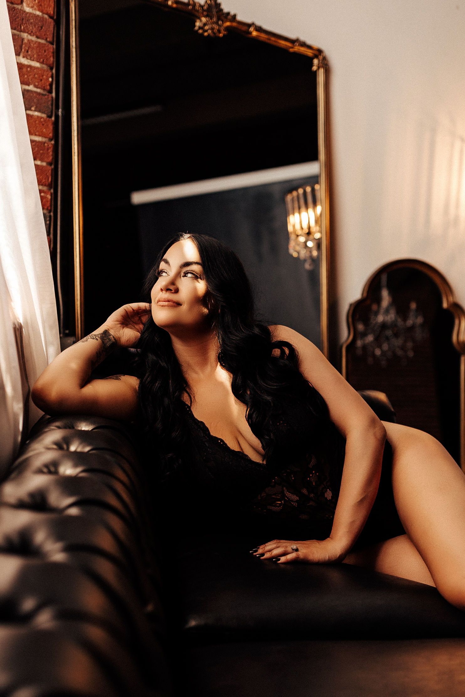 A woman wearing dark lingerie poses on a leather couch in a dimly lit room with exposed brick walls and a mirror.