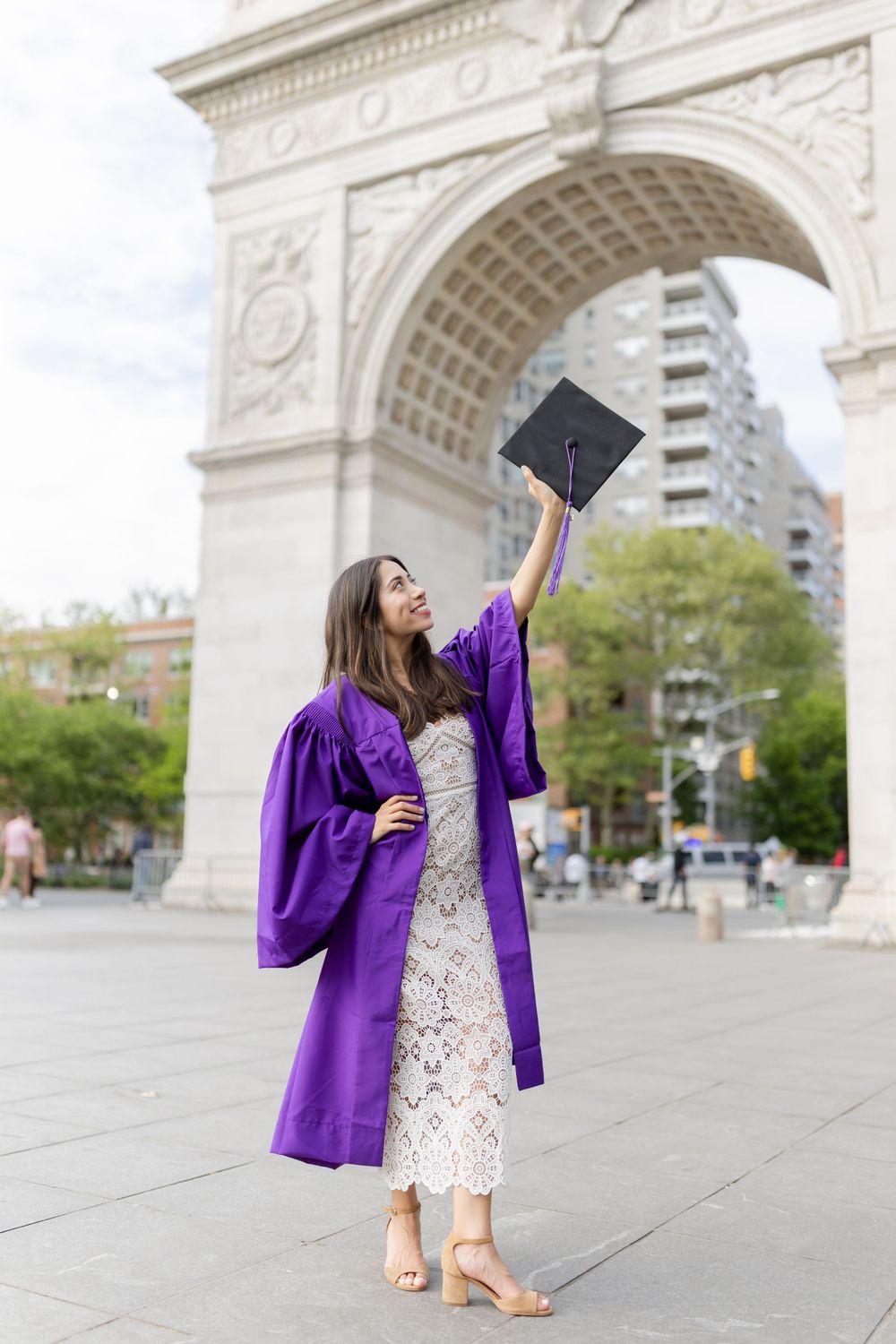 Graduate in purple graduation robe holds up diploma cap in front of Washington Square Arch.