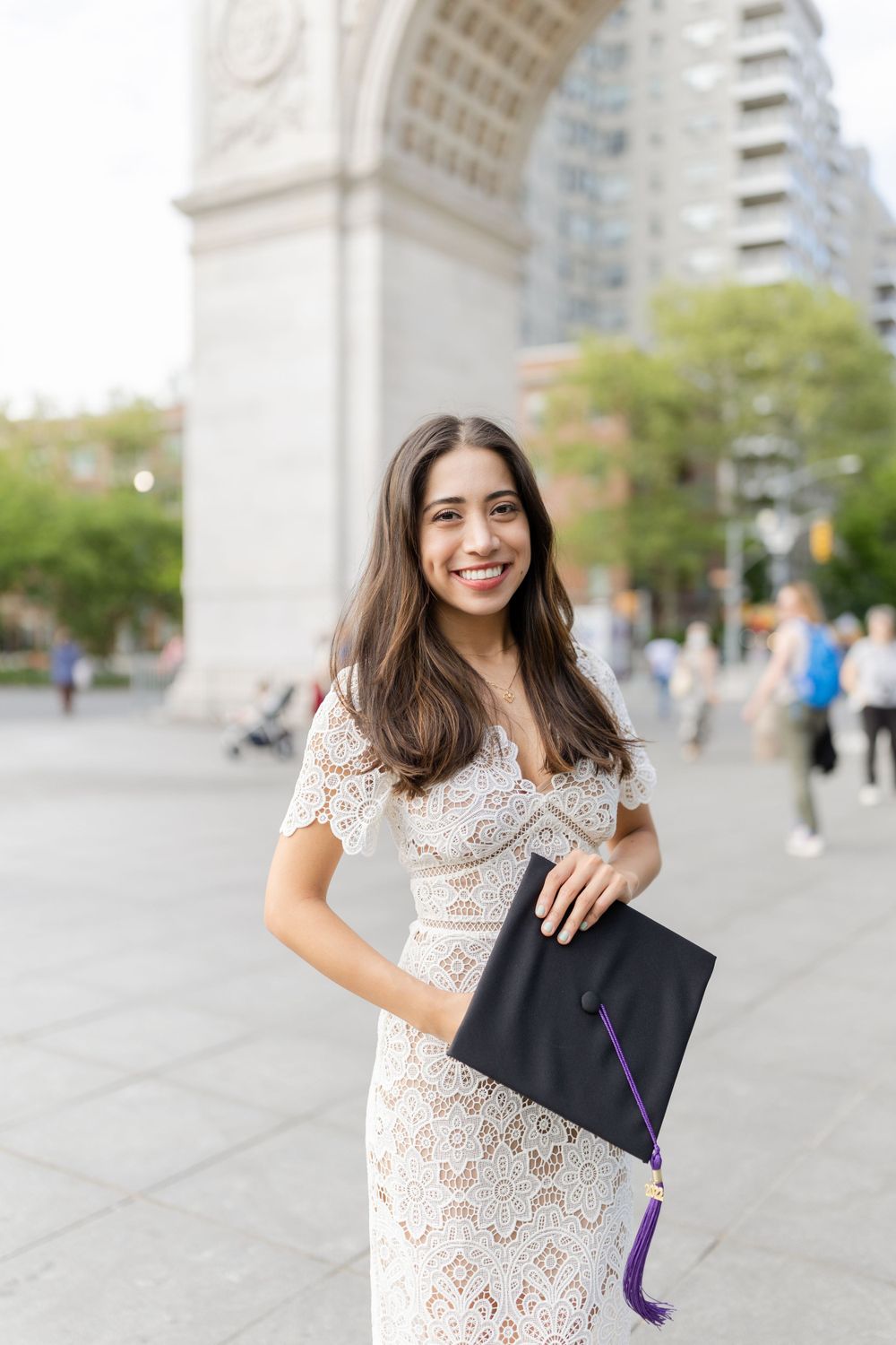 Graduate in white lace dress poses with diploma at Washington Square Park.