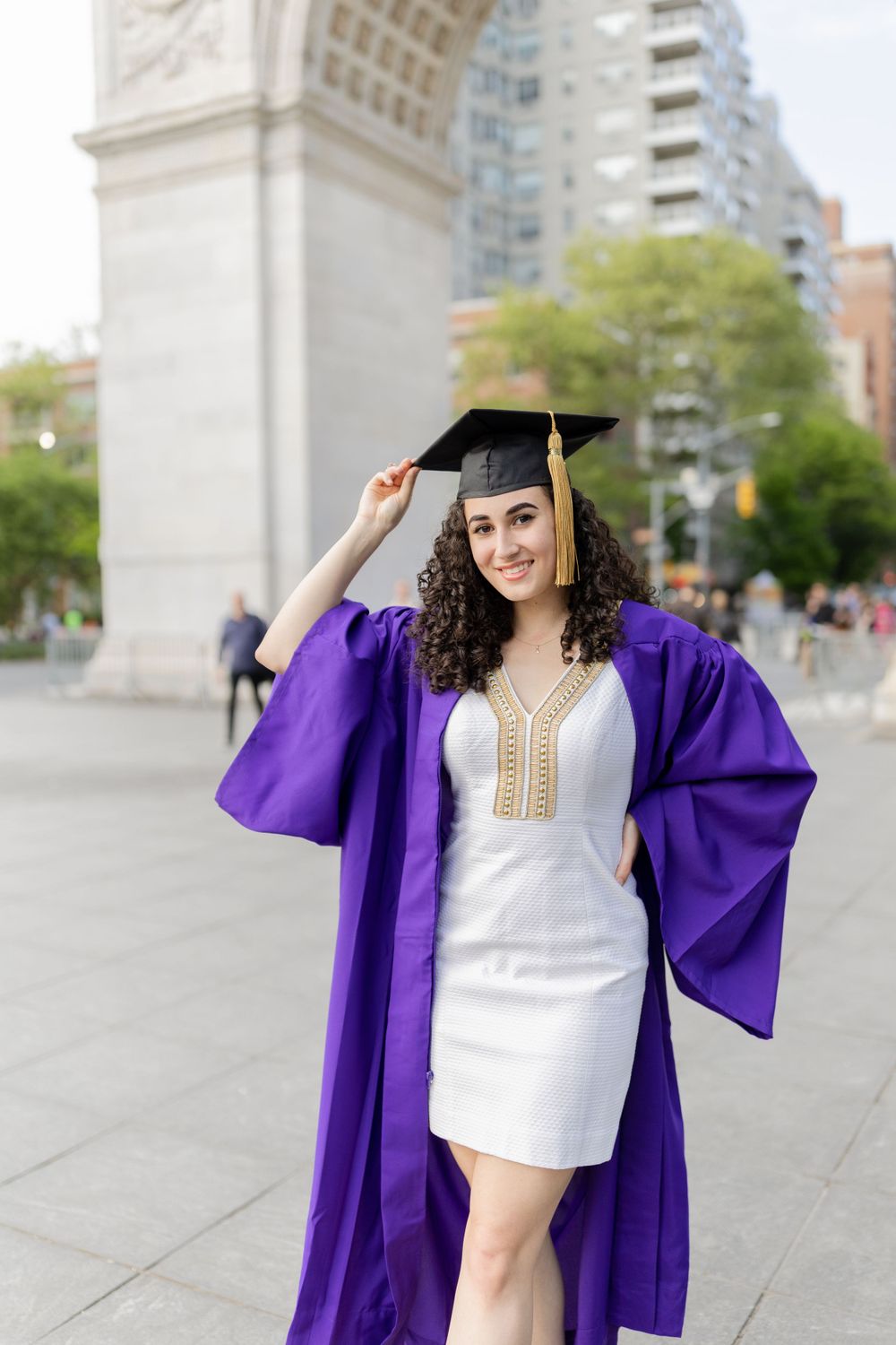 Graduate in purple commencement robe poses with diploma near Washington Square Arch.