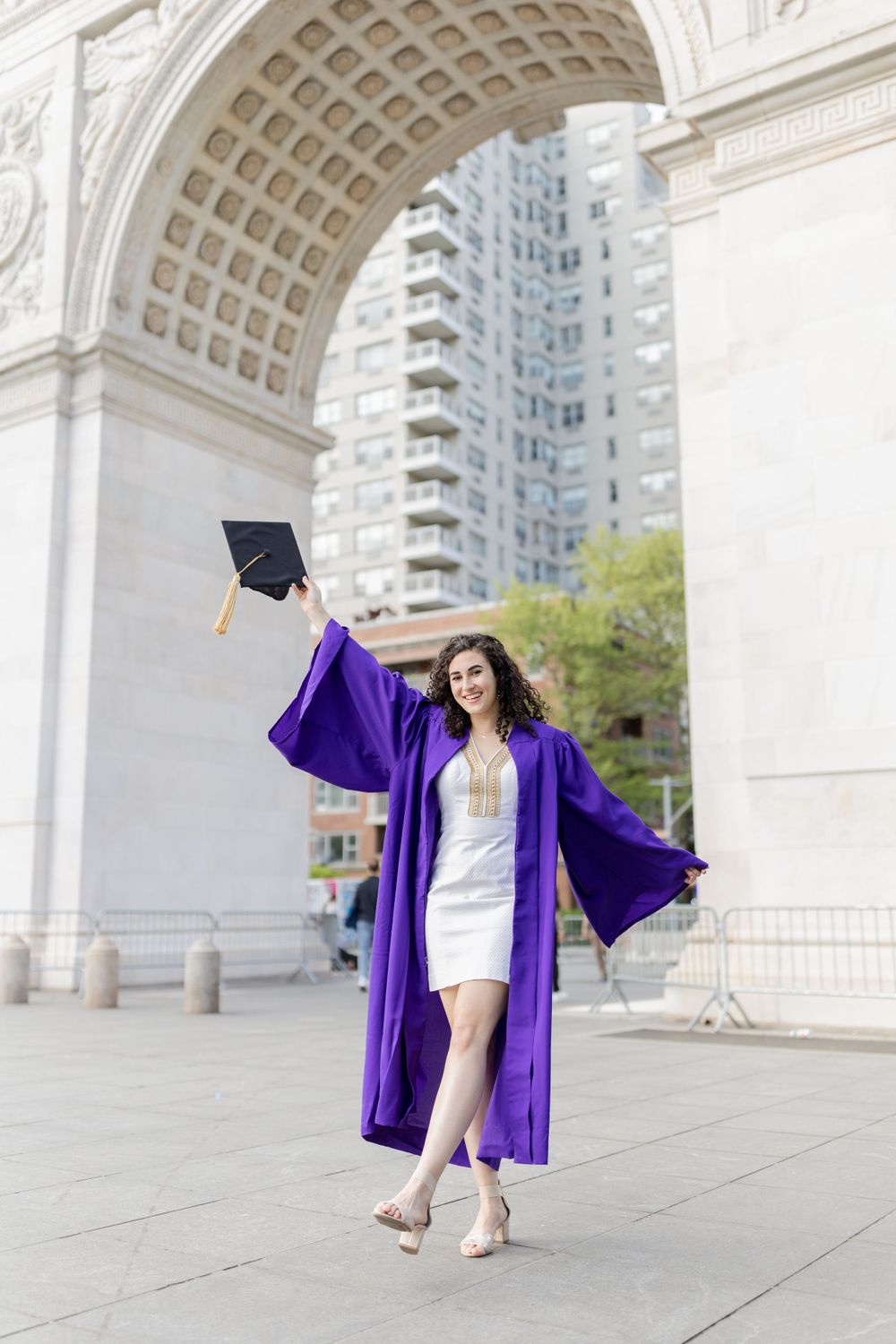Graduate in purple robe twirls while holding cap near Washington Square Arch.