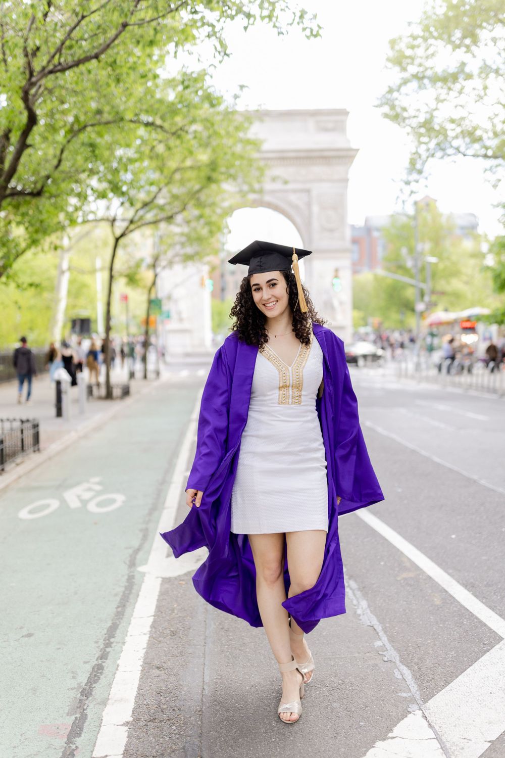 Graduate in white dress and purple graduation gown poses on NYC street with arch backdrop.