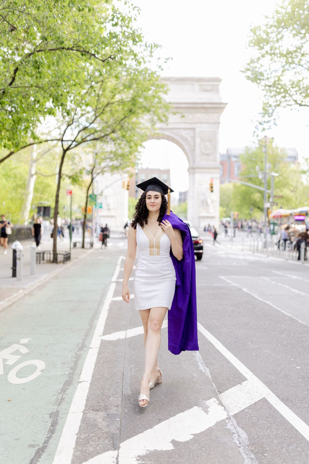 Graduate in purple cap and gown walks along street with Washington Square Arch behind.