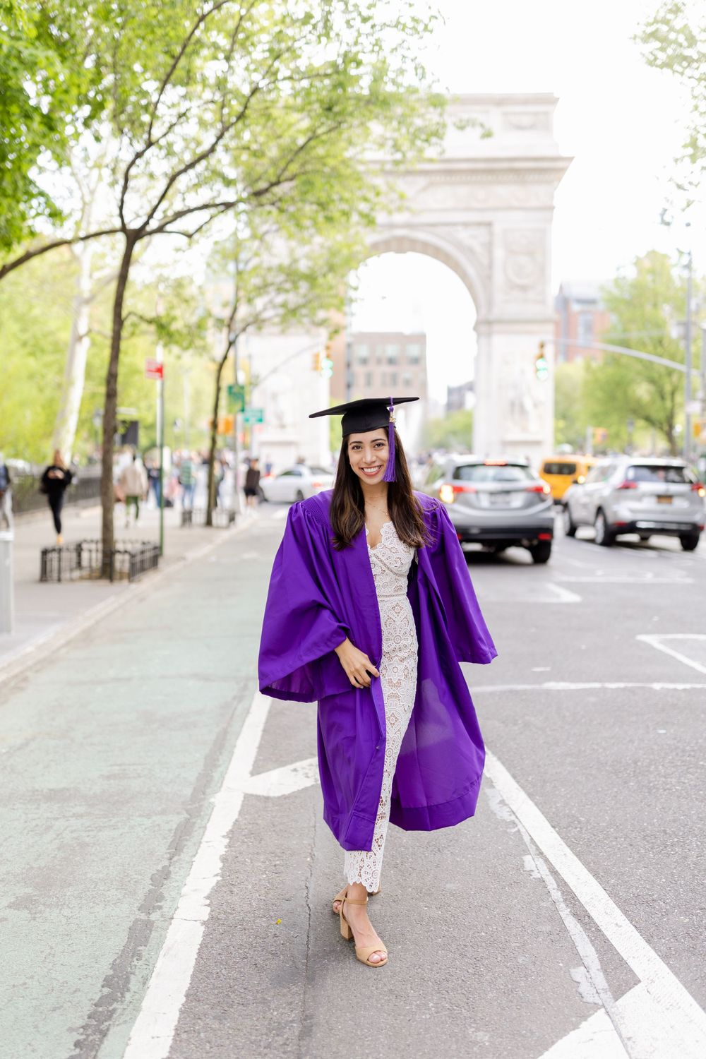 Graduate wearing purple graduation regalia walks down street near iconic Washington Square Arch.