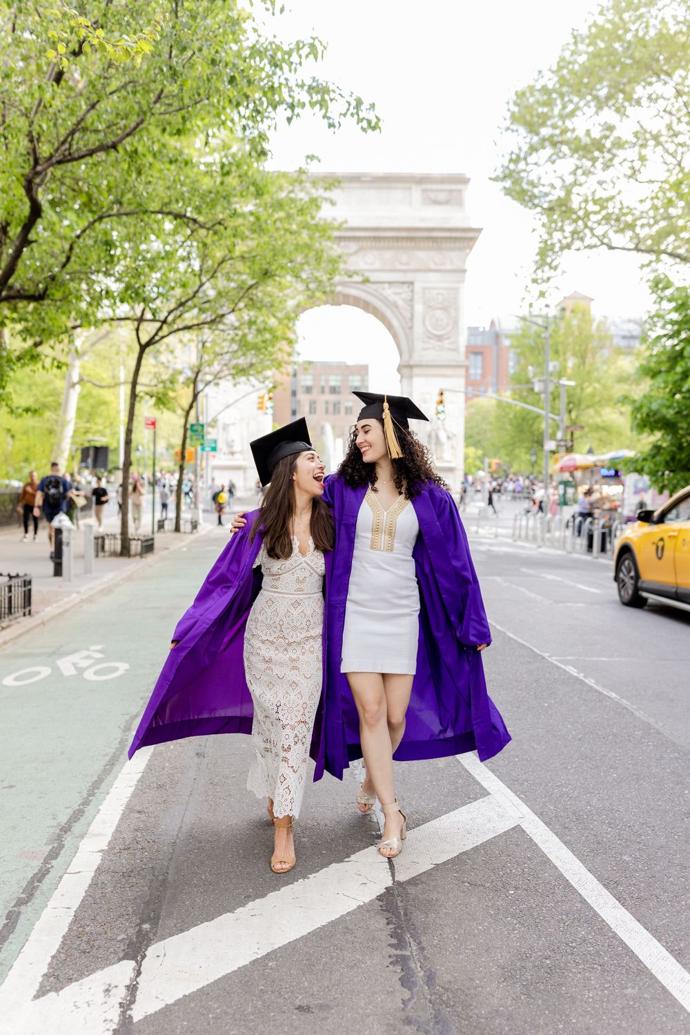 Two graduates in matching purple gowns dance on street near Washington Square Arch.