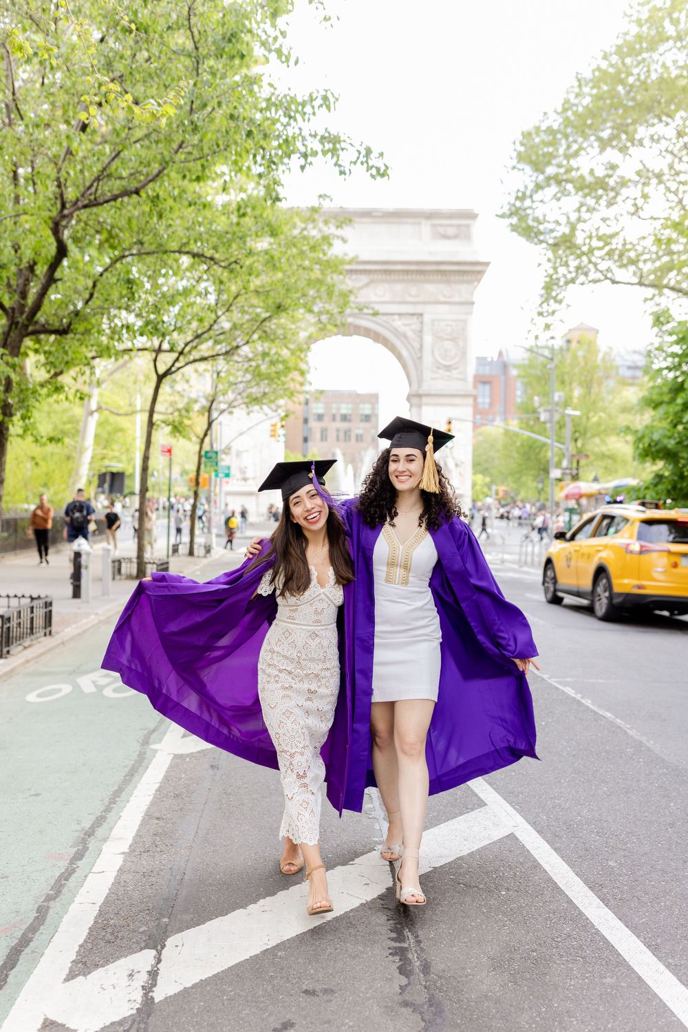 Two graduates in purple regalia pose together on NYC street with arch in background.