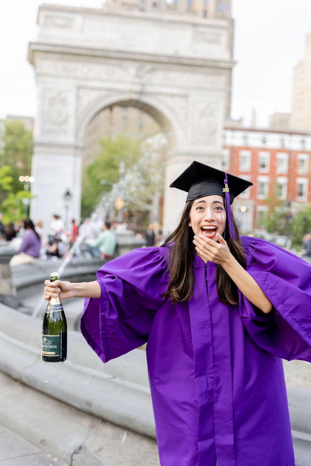 Graduate celebrates with champagne in purple graduation gown in front of Washington Square Arch.
