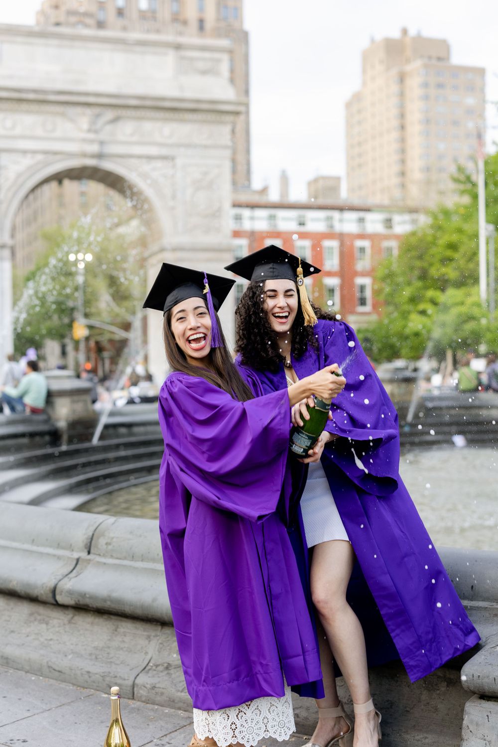 Graduates laughing and celebrating in purple robes by Washington Square Park fountain.