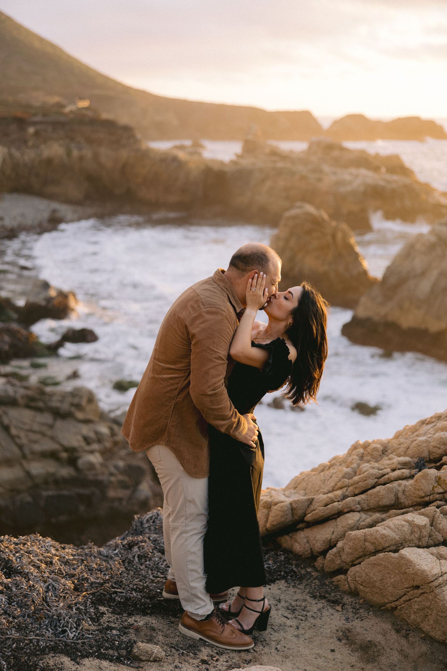A romantic sunset moment shared between two people embracing on rocky coastal cliffs by the ocean.