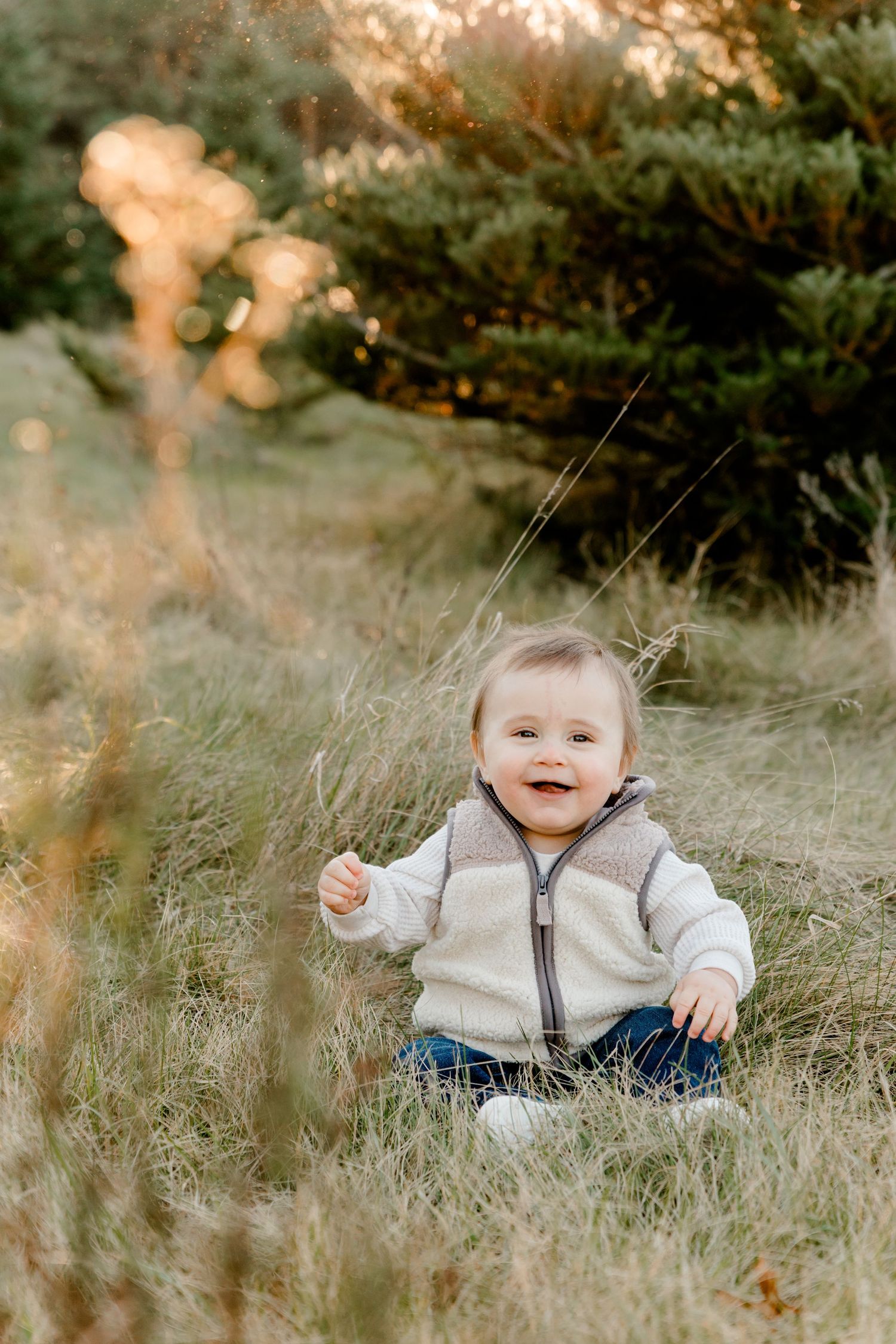Smiling baby in beige zip-up jacket sitting in golden grass near evergreen trees outdoors.