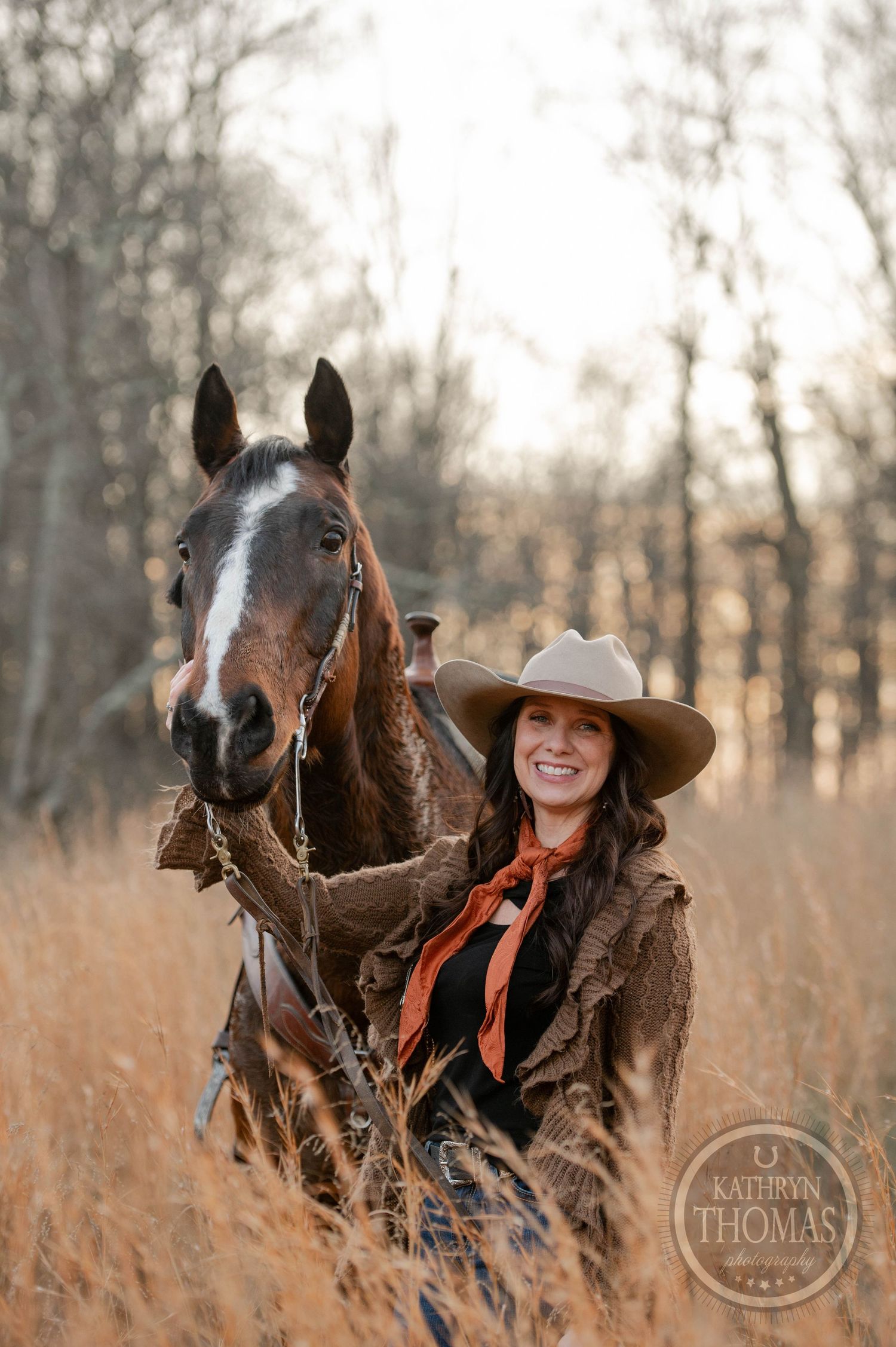A cowgirl in western attire poses with her dark bay horse in a golden field with bare trees in the background.