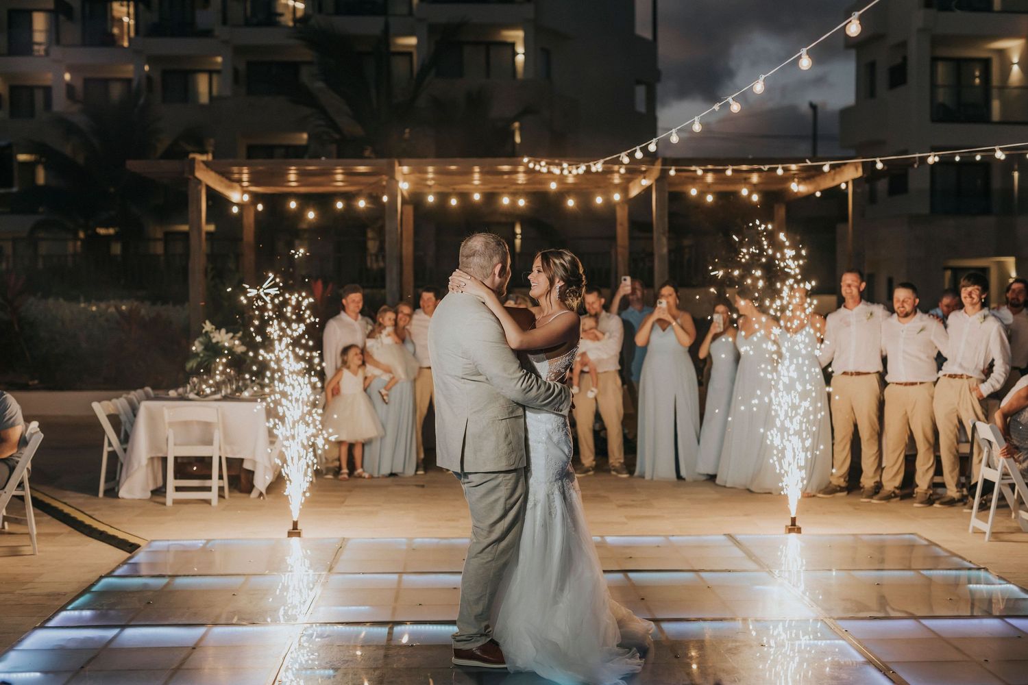 Beautiful outdoor evening wedding reception with couple dancing on LED-lit dance floor under string lights and sparklers.