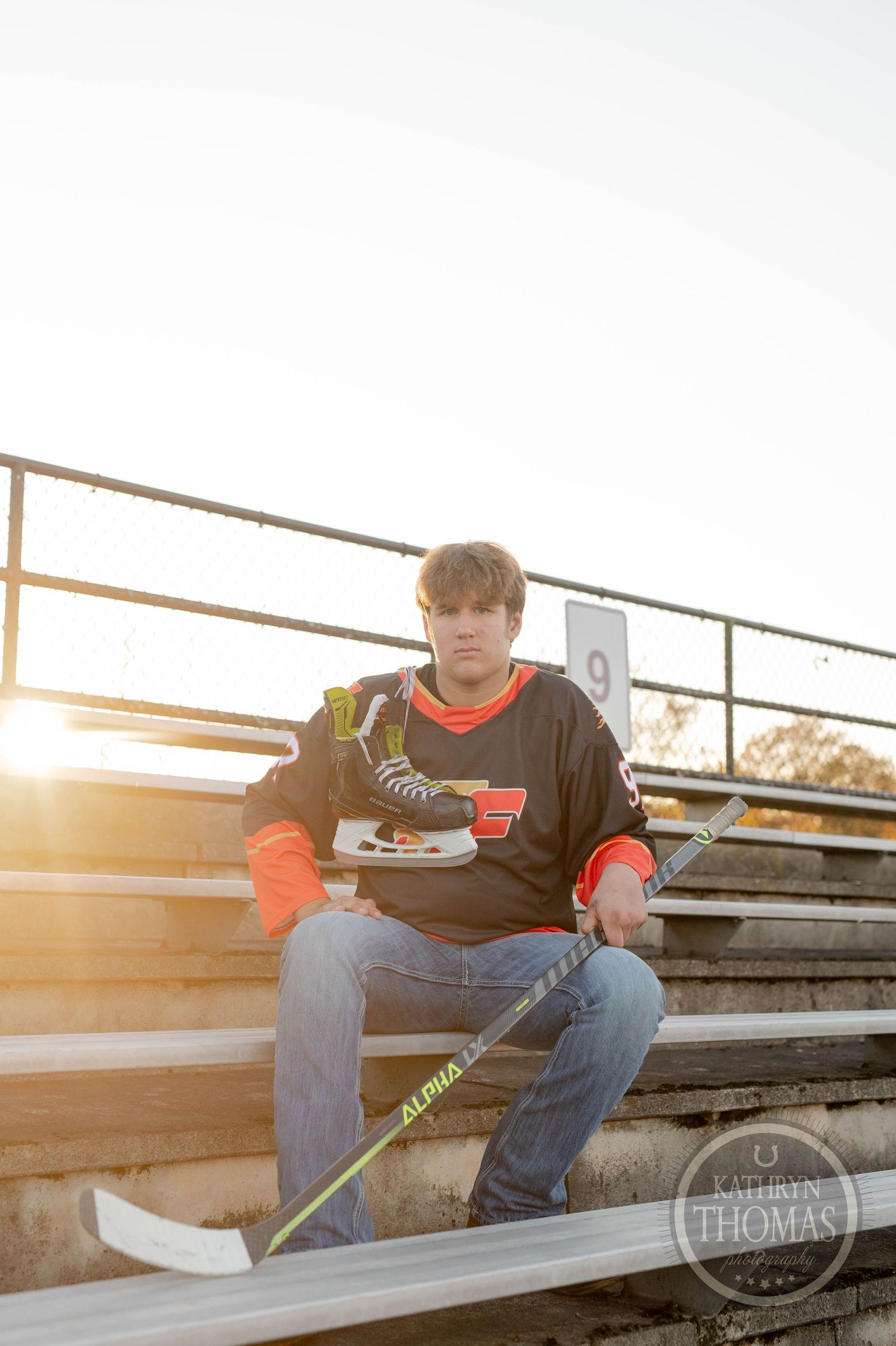 Student athlete poses with hockey equipment on stadium bleachers during golden hour sunset.