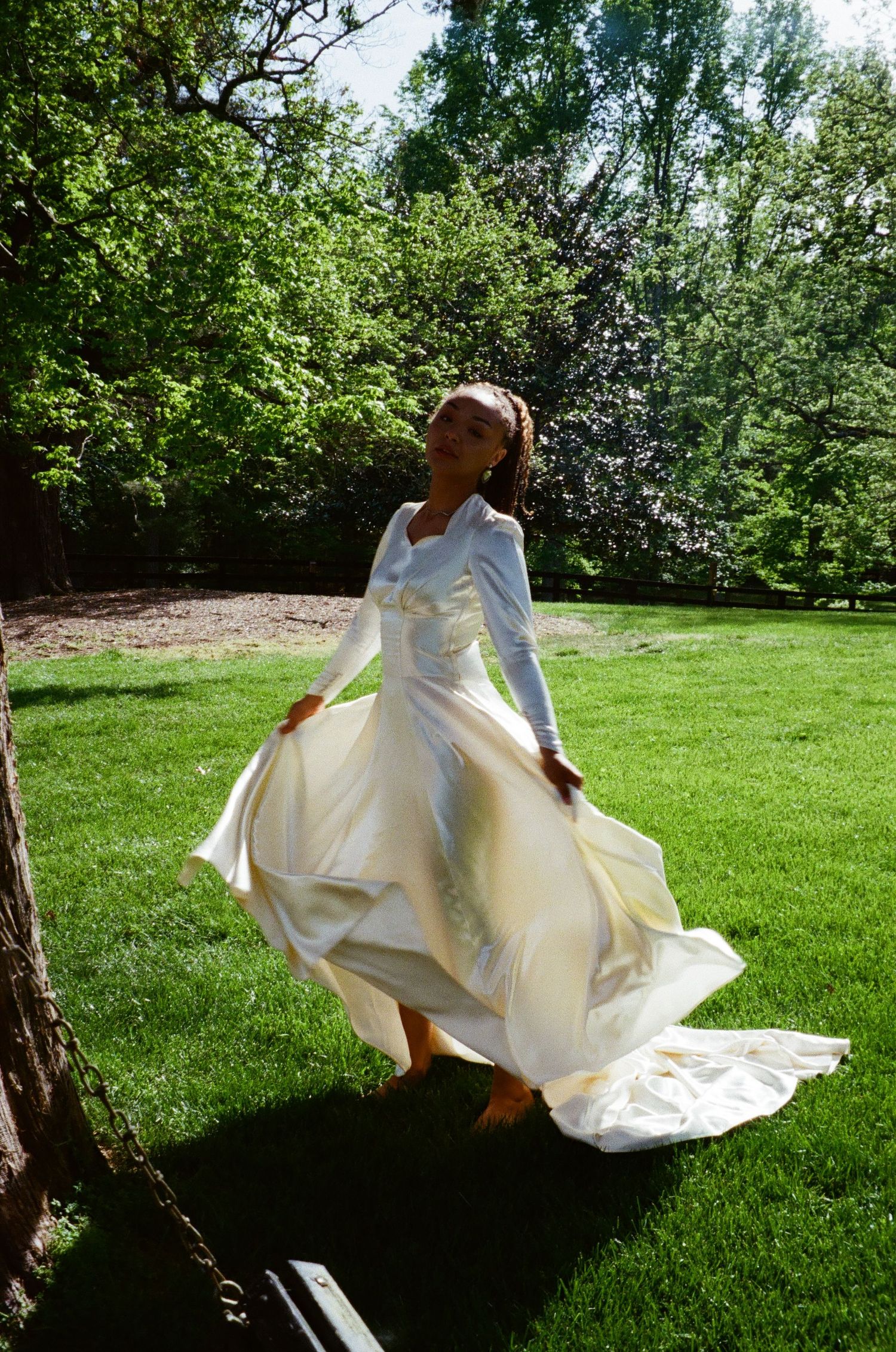 A bride in a flowing white wedding dress twirls gracefully on a sunlit lawn surrounded by lush green trees.