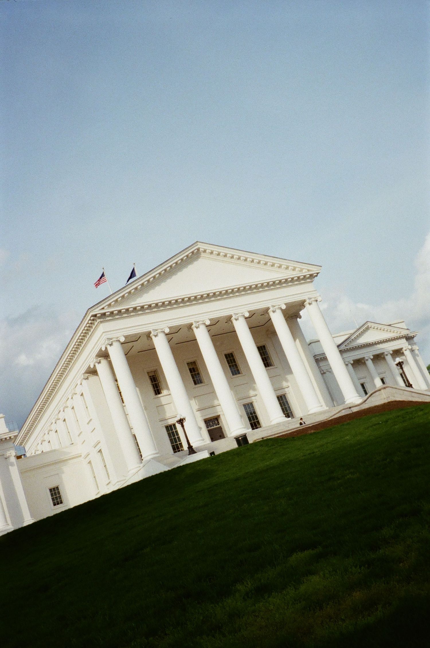 The Virginia State Capitol building with its grand white columns and triangular pediment sits atop a green grassy hill.