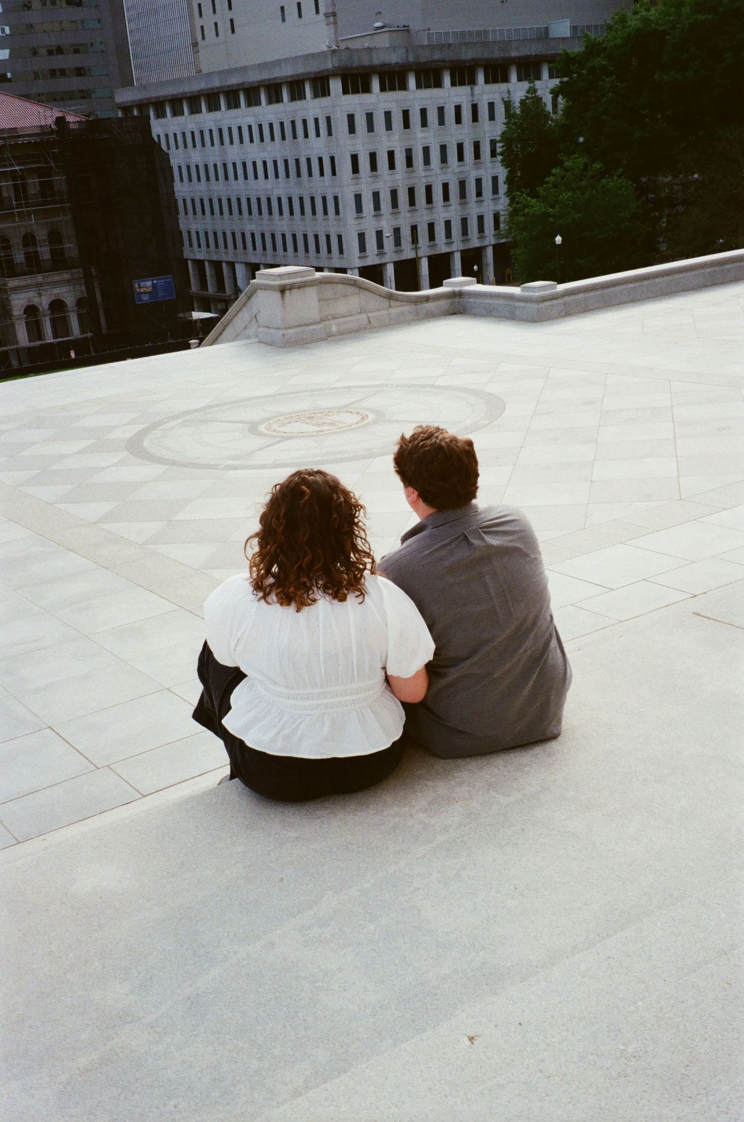 Two people sit close together on a white concrete surface in an urban setting with buildings in the background.