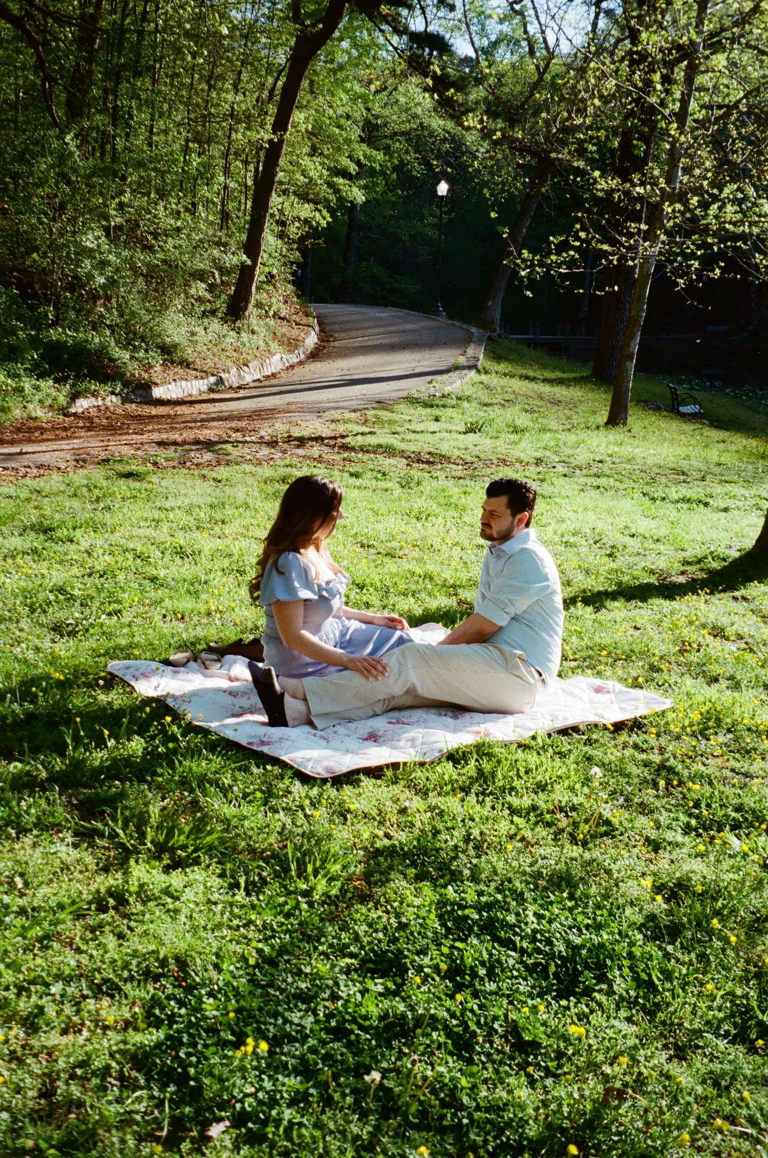 Two people sit on a picnic blanket having a relaxed conversation in a sunny park with winding path and green surroundings.