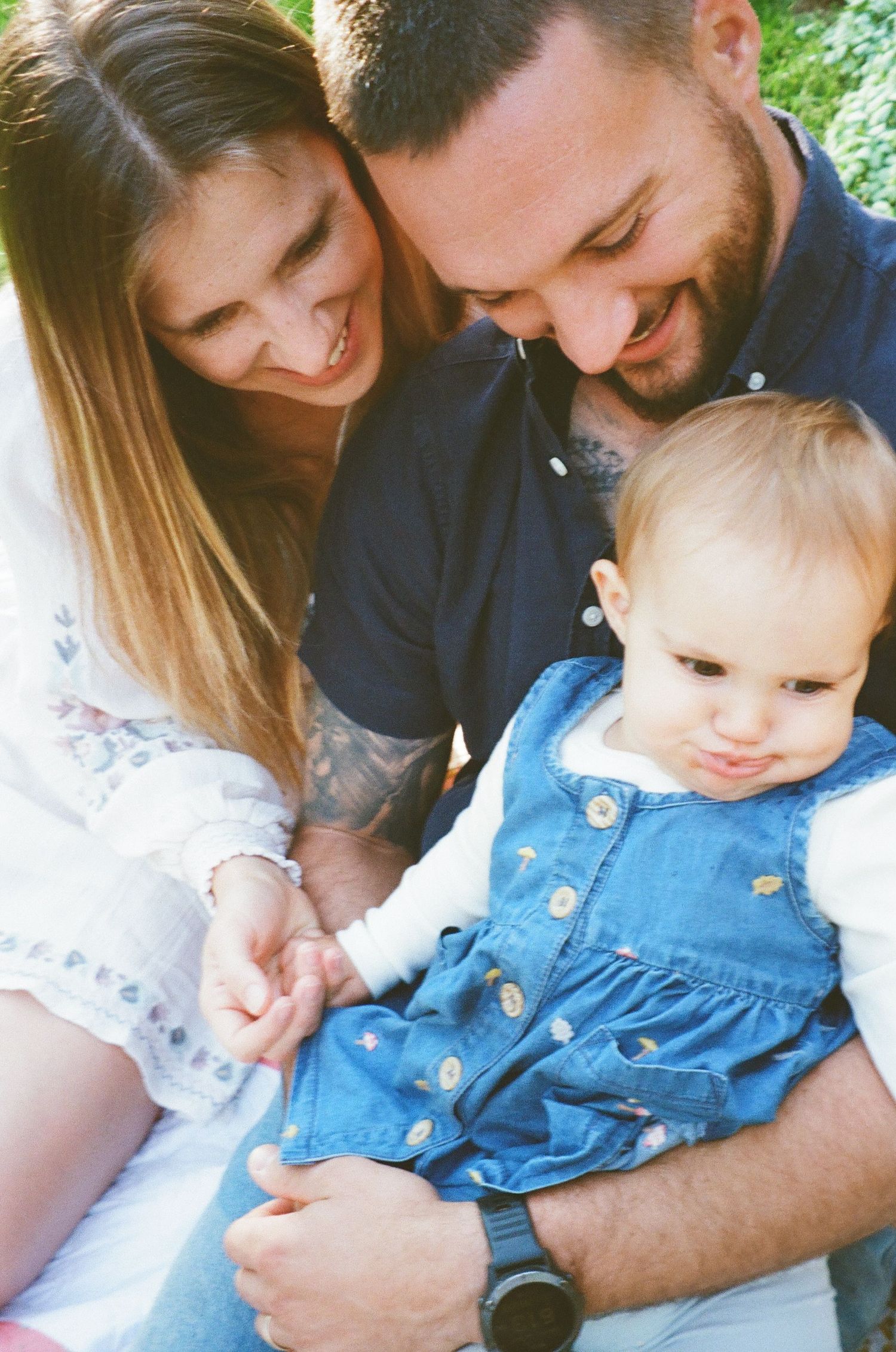 Family shares tender moments with baby wearing denim overalls in outdoor setting.
