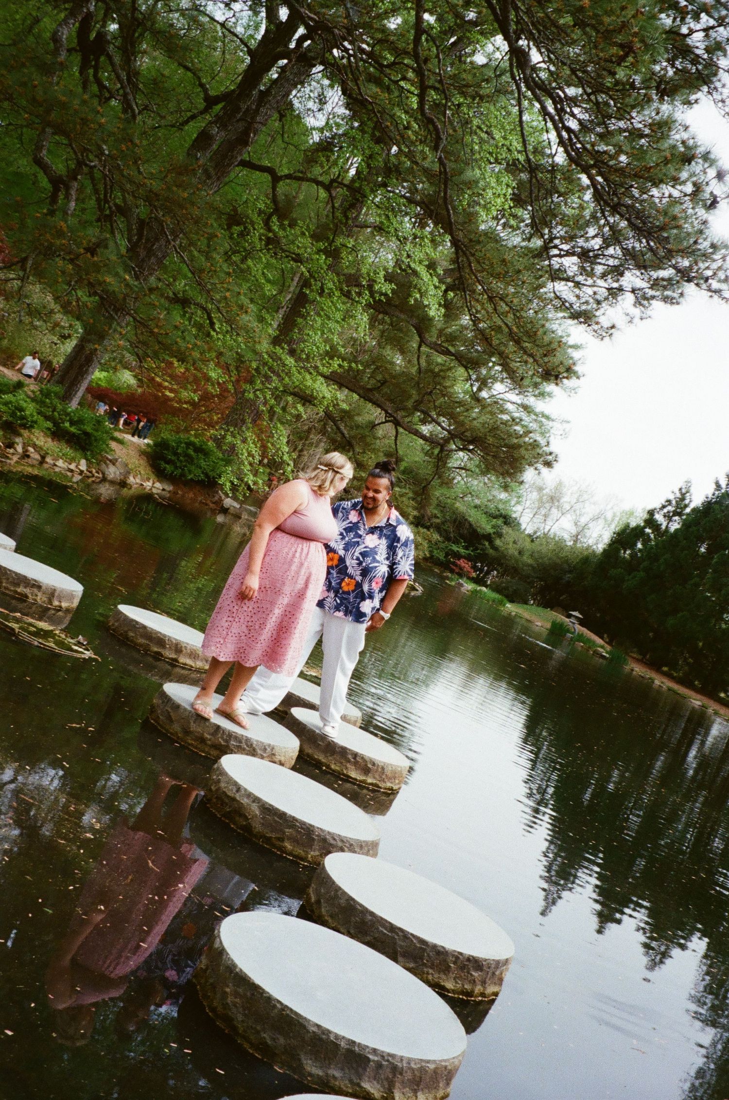 A couple stands together on stepping stones across a tranquil pond surrounded by lush greenery on a summer day.