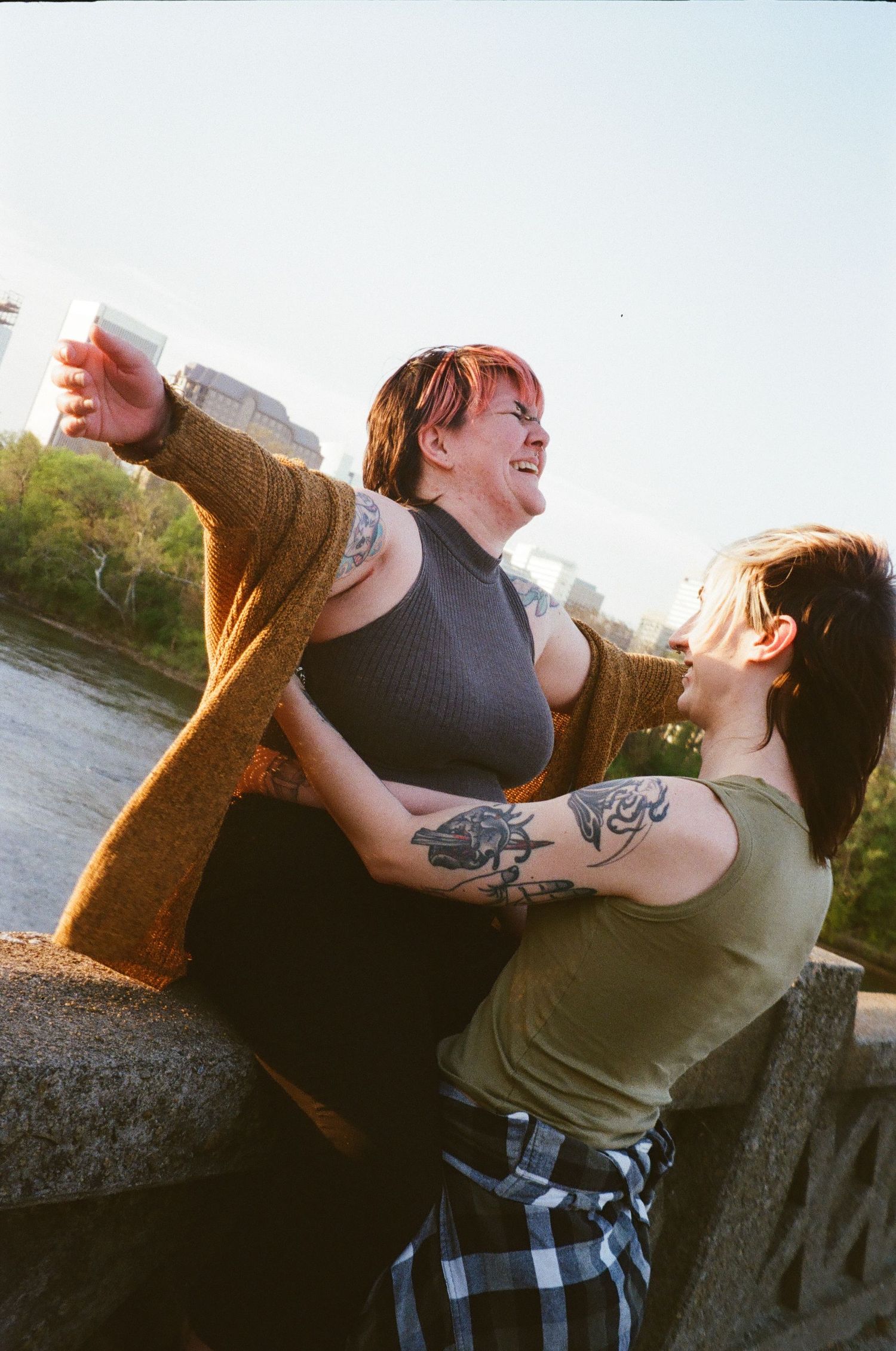 Two tattooed women laughing and embracing outdoors near a river on a sunny day.