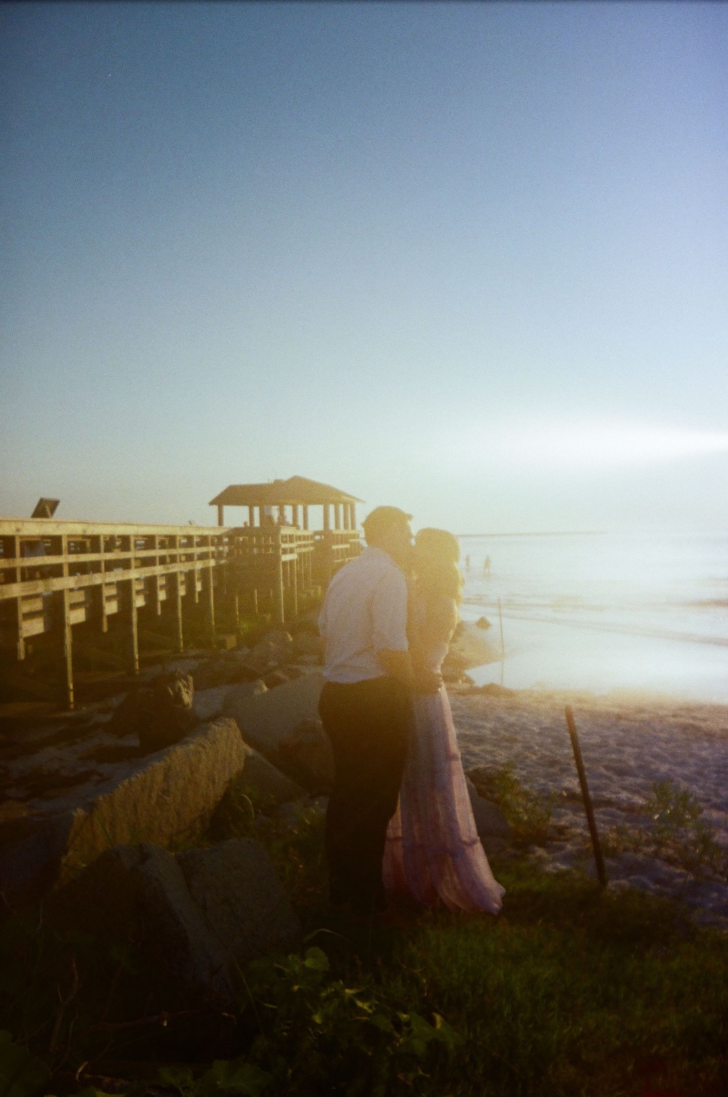 A couple silhouetted at sunset near a wooden pier, with a gazebo visible in the warm hazy background.