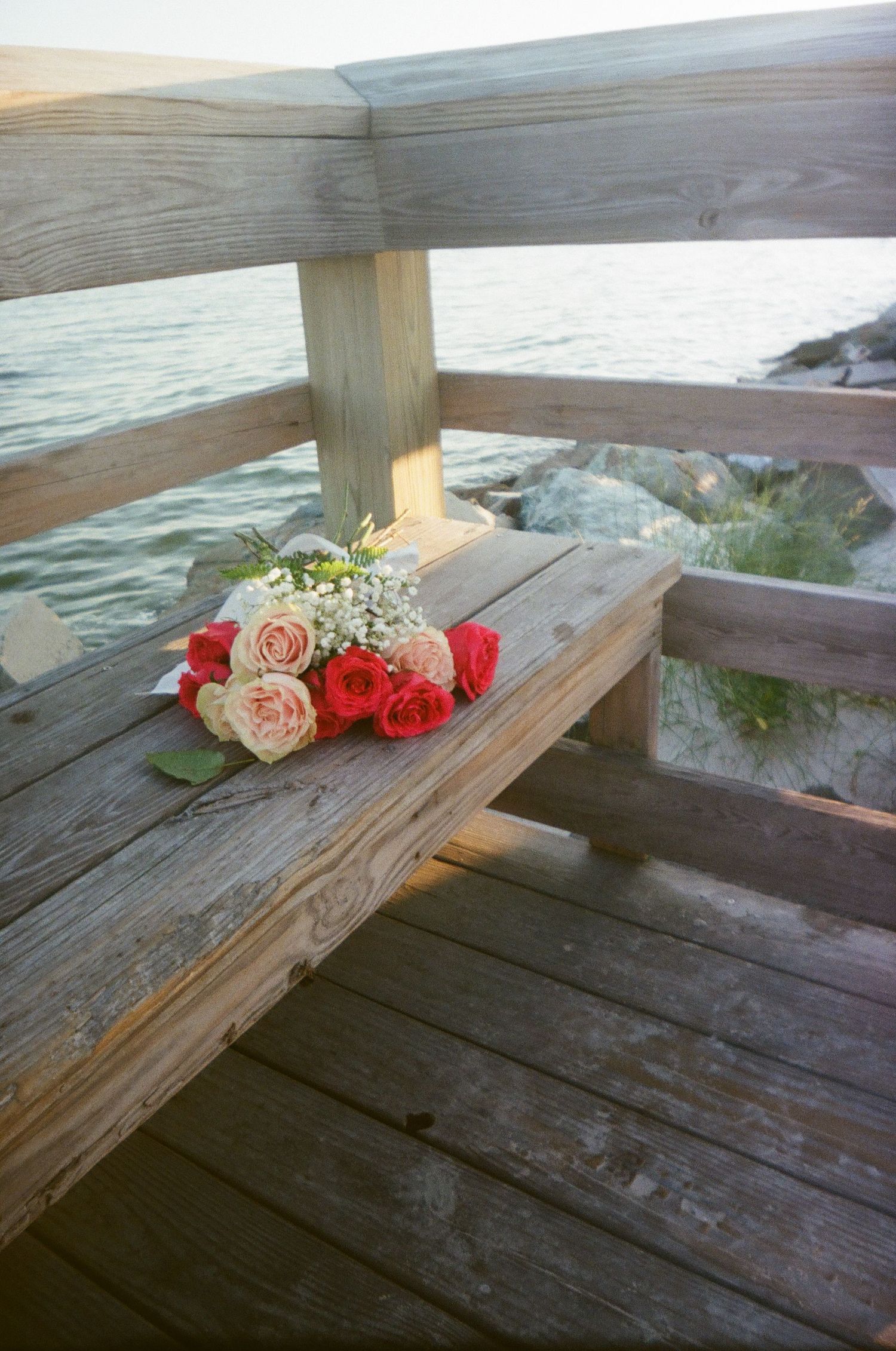 A bouquet of pink and red roses rests on a wooden deck railing overlooking the ocean at sunset.