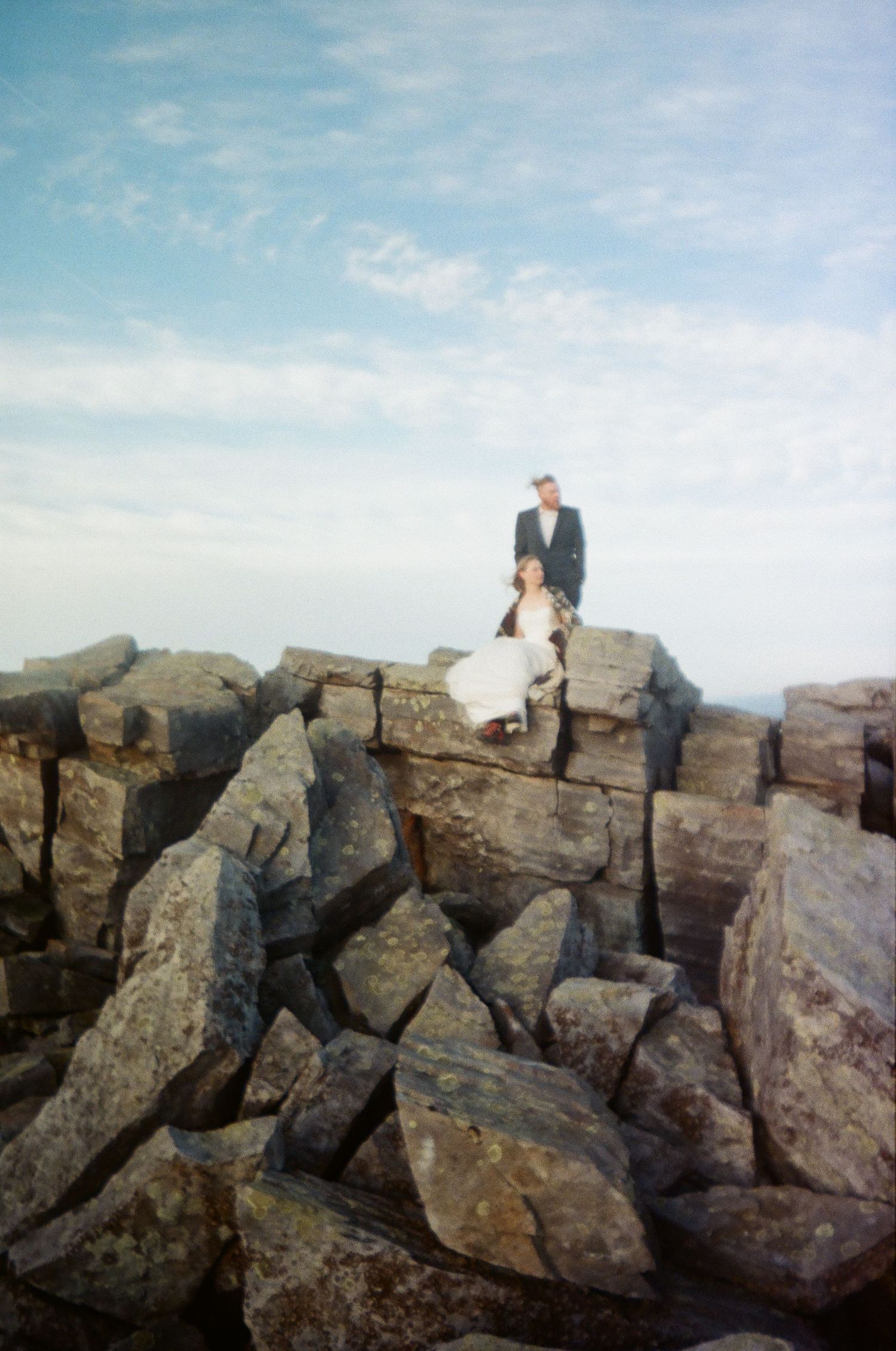 A couple stands together on a dramatic rocky outcrop against a soft blue sky.