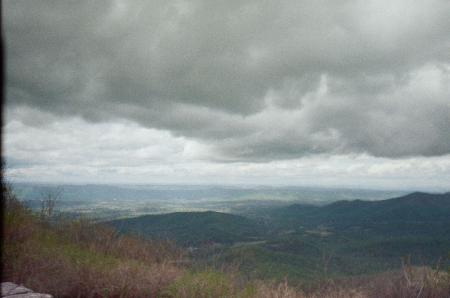 Dramatic stormy clouds loom over a mountainous landscape with rolling hills stretching into the distant horizon.
