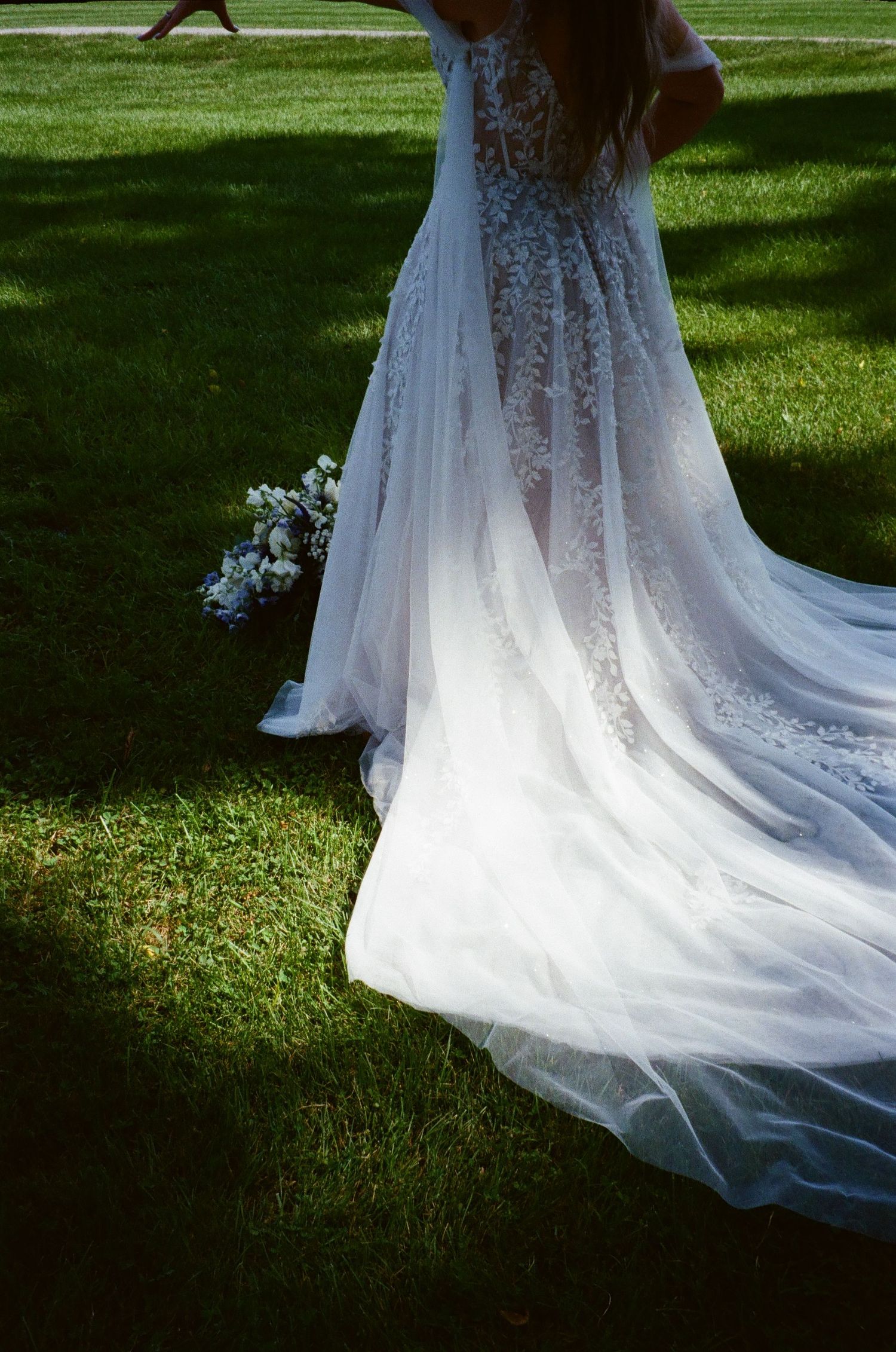 Elegant lace wedding dress with flowing train spreads across green lawn in outdoor summer setting.