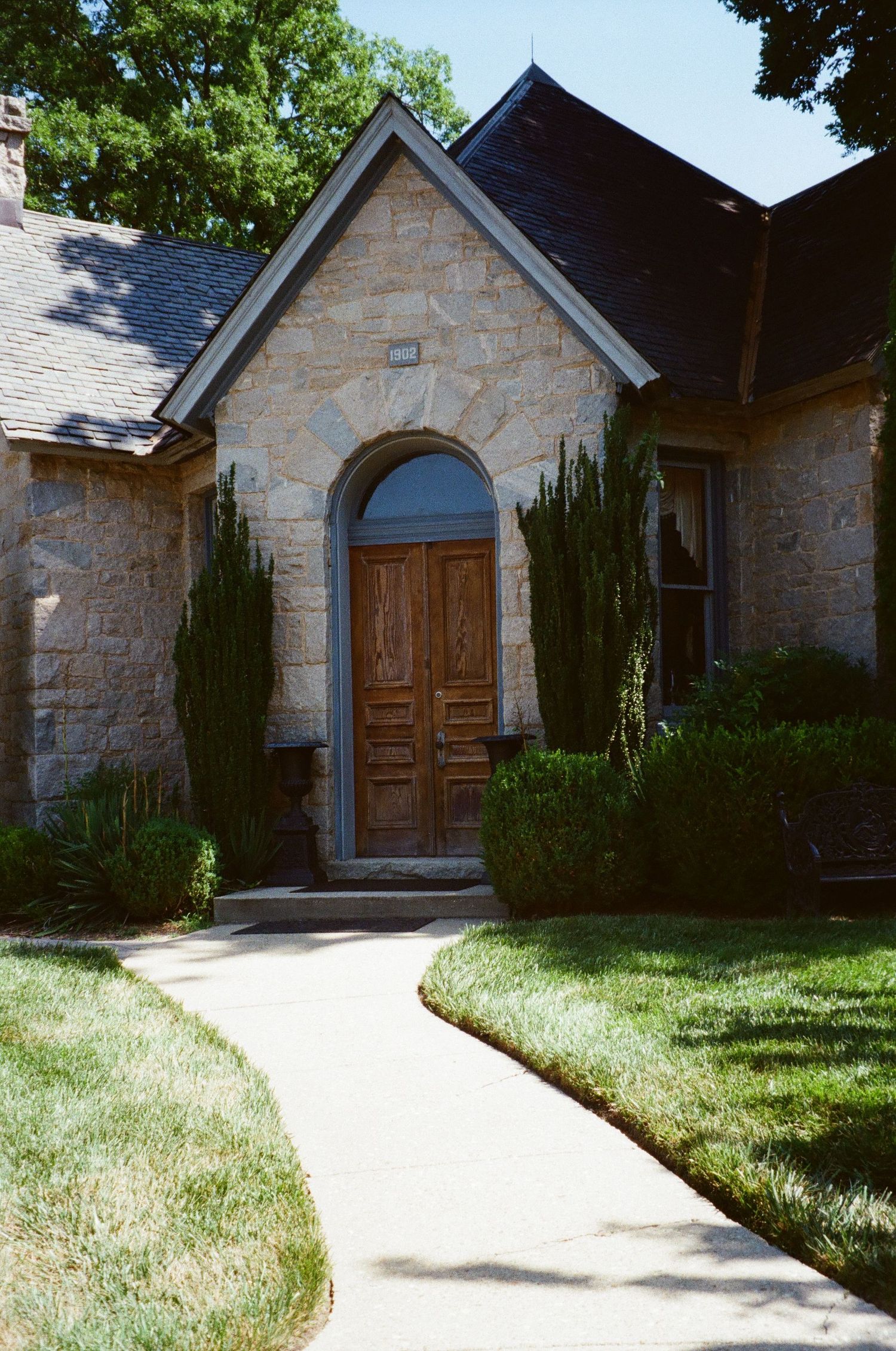 Stone facade cottage-style home with wooden doors and cypress trees lining a curved concrete pathway.