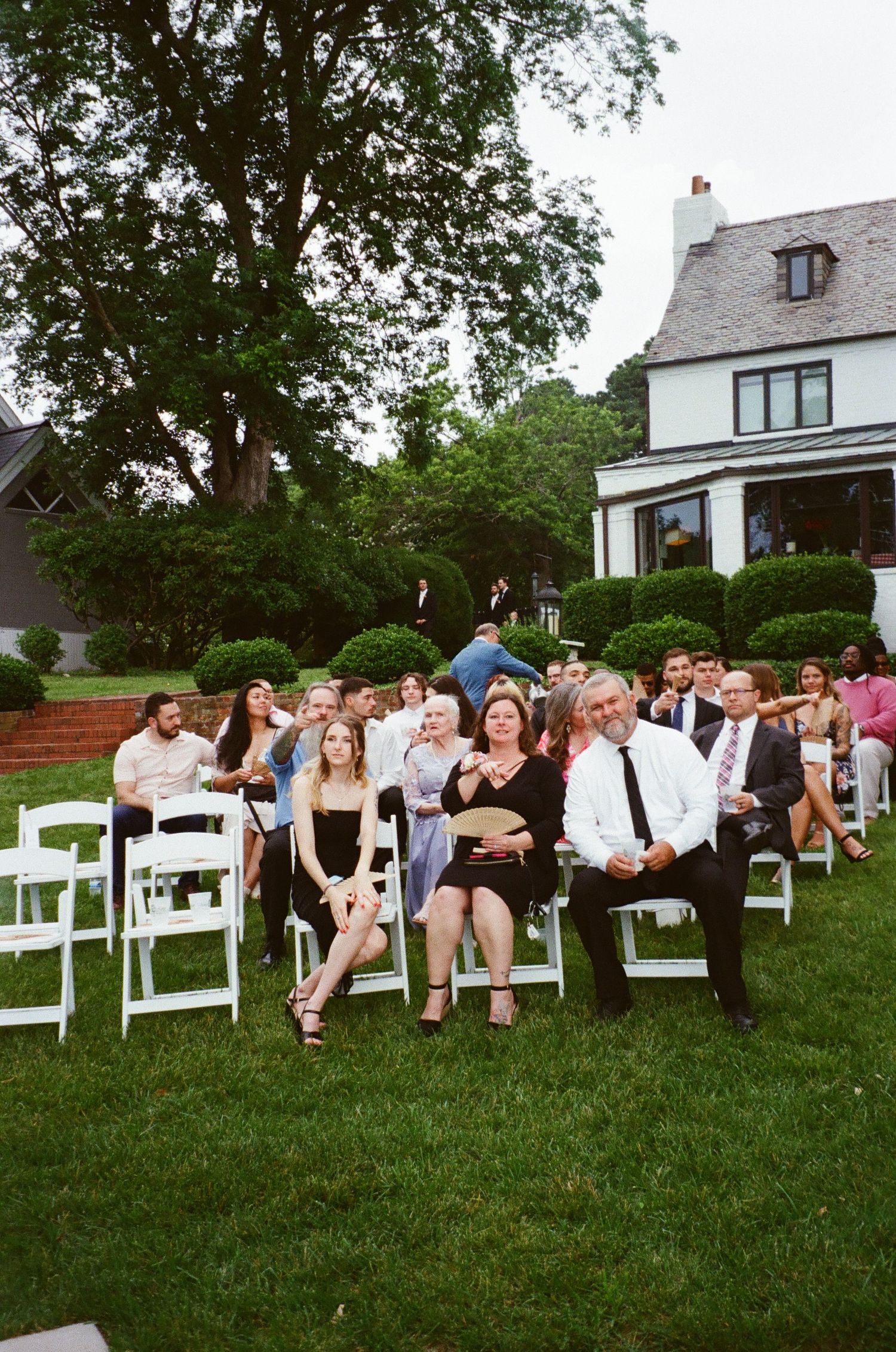 Wedding guests sitting in white chairs on a green lawn in front of a charming white house during an outdoor ceremony.