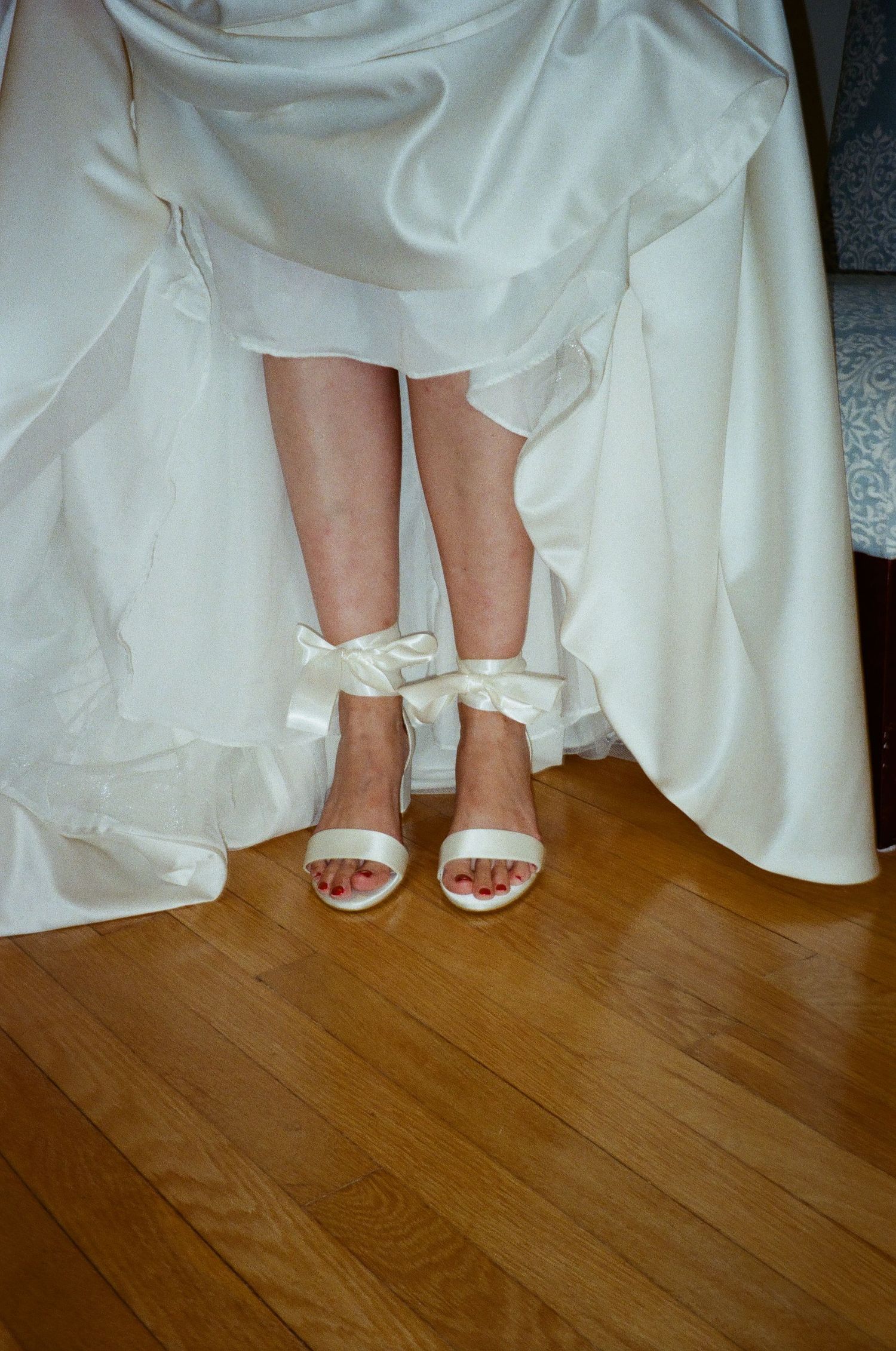 Close-up of bride's feet in white sandals with bow details peeking out from under a flowing wedding dress.